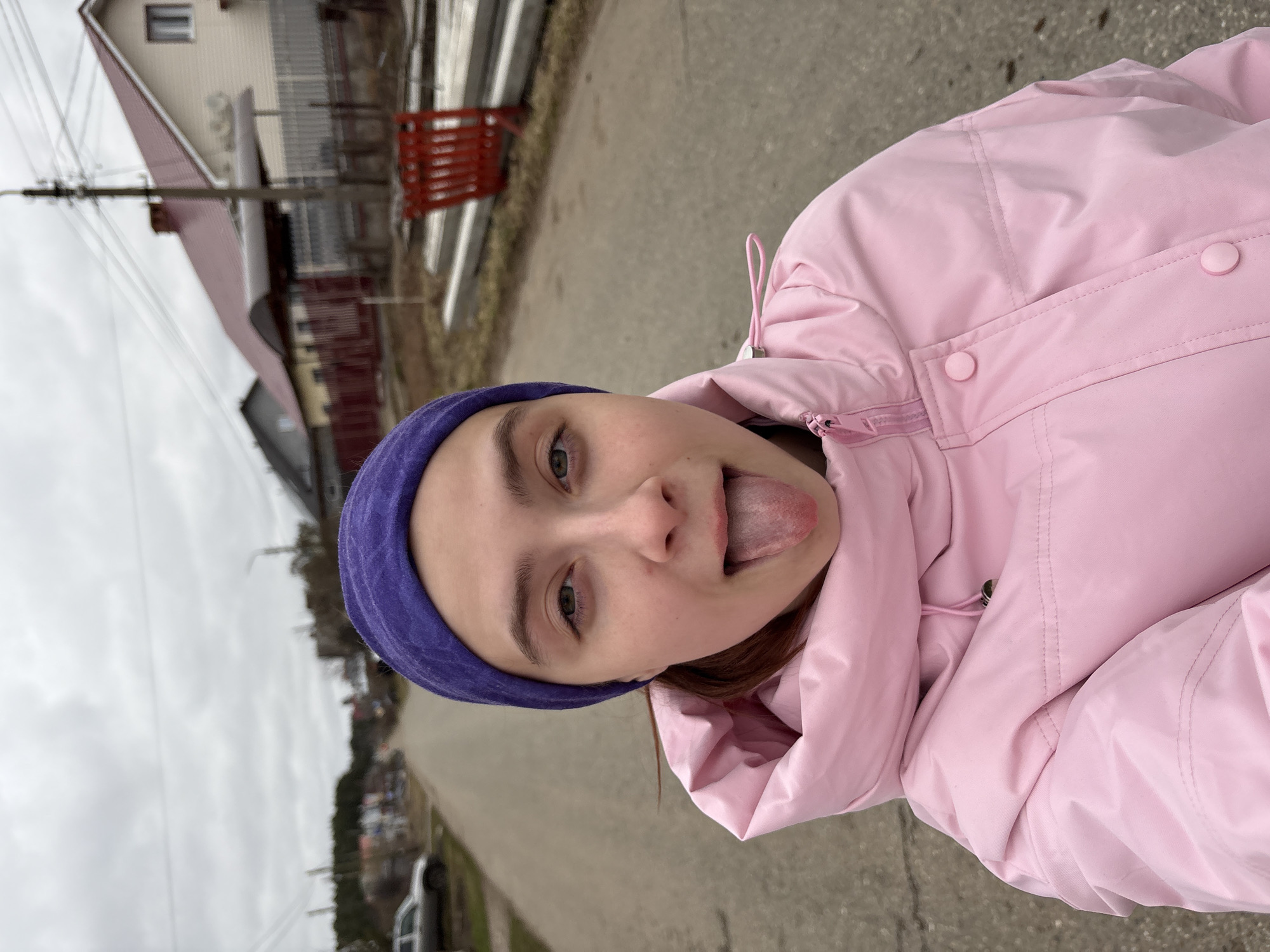 A young person is captured in a casual outdoor portrait, rotated on its side. The subject, who appears female, is wearing a bright pink puffy jacket and a deep purple beanie. She looks directly at the camera with a playful expression, sticking her tongue out. Her eyes are a light shade, possibly green or hazel. The pink jacket features a visible zipper, seams, and two light pink buttons on the front panel.
The background depicts a residential street scene, likely in a rural or suburban part of Прикамский, Russia. To the left, behind the subject, several houses are visible, including one with a reddish-brown roof and light-colored siding. Utility poles with power lines crisscross against an overcast, grey sky. A road, appearing to be paved or dirt, extends into the distance, flanked by some vegetation and what might be a parked vehicle further down. The weather seems cool and cloudy, fitting for the warm attire of the subject. The overall impression is one of a candid and lighthearted moment. There is no visible text in the image.