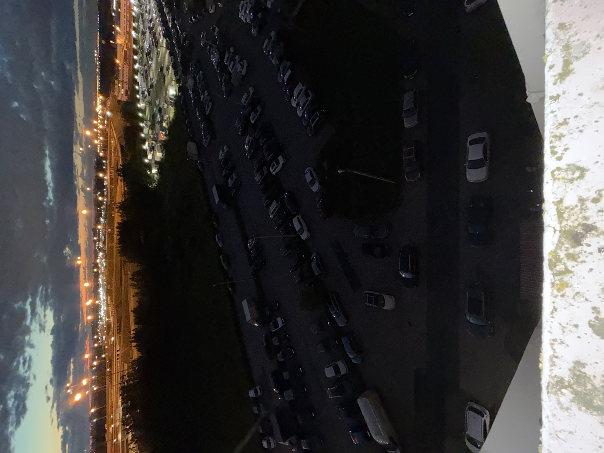 The image, taken at dusk, overlooks a vast expanse of urban landscape. In the foreground, a portion of a white, weathered railing with green moss visible suggests a vantage point from an elevated structure, possibly a balcony or rooftop.
Below, a large, dimly lit parking lot stretches across the frame, filled with numerous cars of various shapes and colors, predominantly dark hues. Several vans are also visible, one white and one light-colored, parked amidst the cars. The parking lot appears to be a central feature of the scene.
Beyond the parking lot, a dense line of dark trees forms a natural border. Further in the distance, the city awakens with a multitude of artificial lights. Streetlights illuminate roadways and the general urban sprawl, creating a warm, glowing haze against the darkening sky. The sky itself is a dramatic canvas of deep blues and grays, with streaks of orange and yellow from the setting sun peeking through scattered clouds. The overall impression is that of a city transitioning from day to night, with the bustling activity of vehicles in the foreground and the city's illuminated network stretching into the horizon.