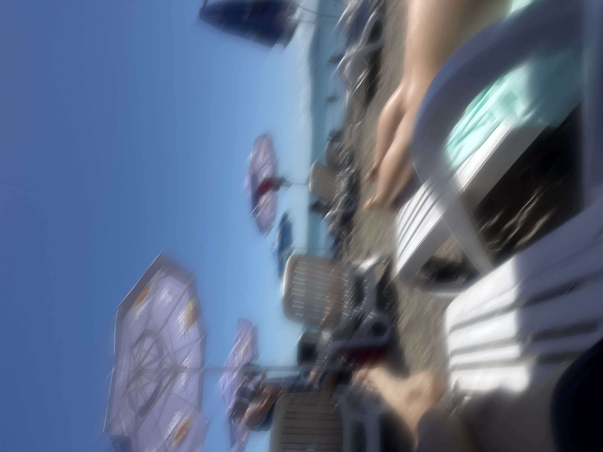 The image shows a beach scene in Burgas, Bulgaria. The shot is taken from a low angle, likely from someone sitting in a white plastic beach chair. Visible are multiple beach umbrellas, a clear blue sky, and a glimpse of the sea in the distance. Other beach chairs are occupied with people. The picture appears blurry, suggesting movement.