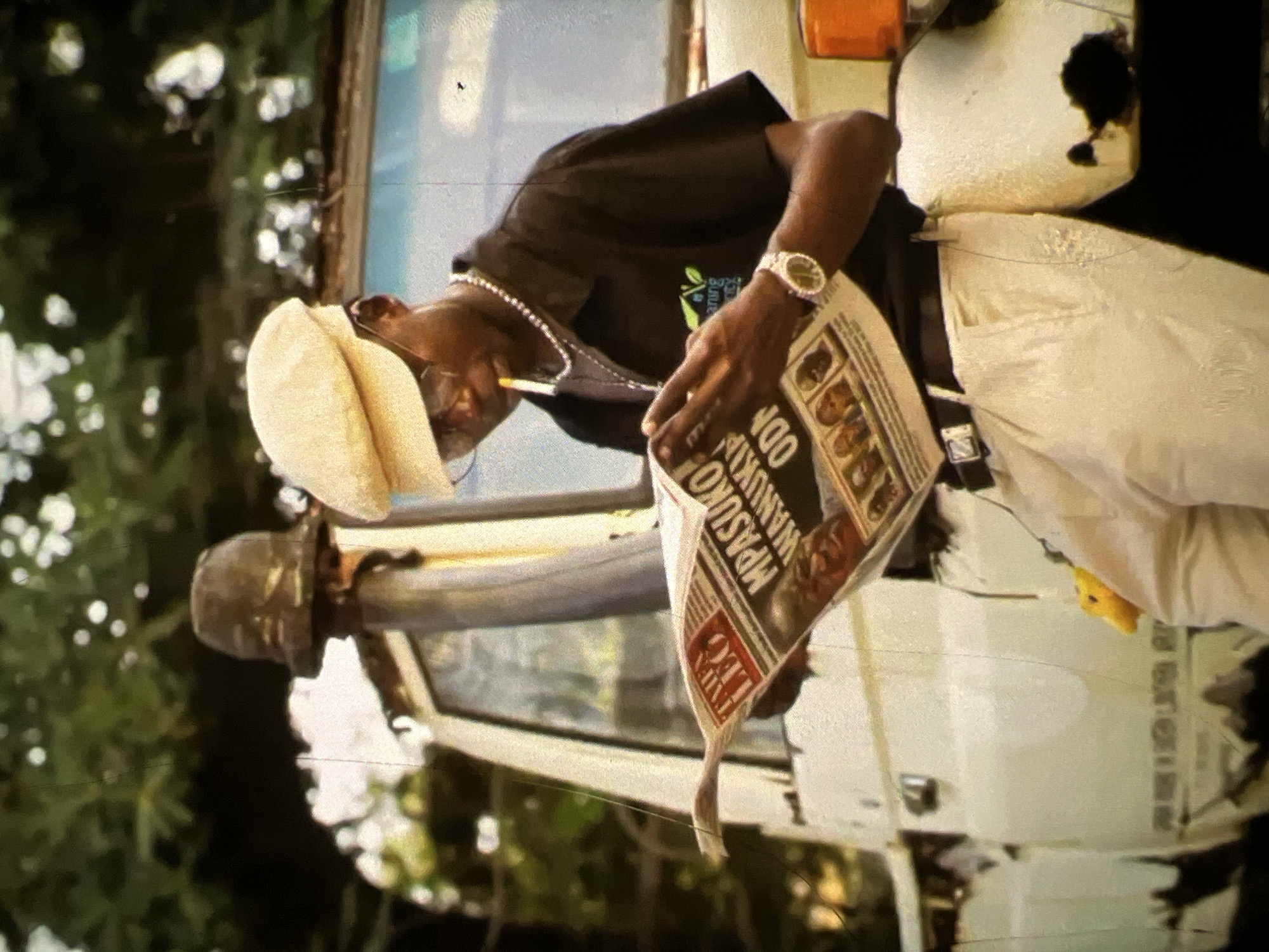 A young Black man stands outdoors, leaning against a white vehicle, reading a newspaper. He wears a light-colored newsboy cap, glasses, a black short-sleeved collared shirt with a green logo on the left chest, a silver chain, light-colored pants, a black belt with a silver buckle, and a silver wristwatch. He is smoking a cigarette. The newspaper he is reading has bold headlines. Behind him, a portion of the vehicle's window is visible, as is some foliage and a dark, old-fashioned helmet perched on a pole. The environment appears to be a street in Lagos, Nigeria, suggested by the style of the vehicle and the general atmosphere. The image has a vintage aesthetic with visible film grain and color shifts.