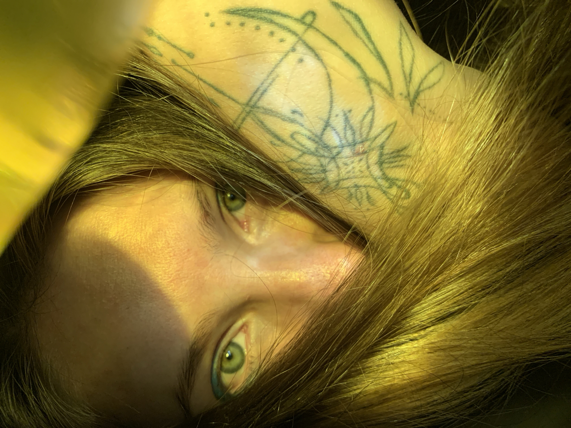 A close-up, overhead shot shows a person's face, mostly obscured by their long, light brown hair. Their eyes are open and looking forward. There is a tattoo on their arm. The lighting is yellow, with a diagonal shadow across their forehead. There are no discernible landmarks or environmental features to suggest a specific location within Chaikovsky, Russia.