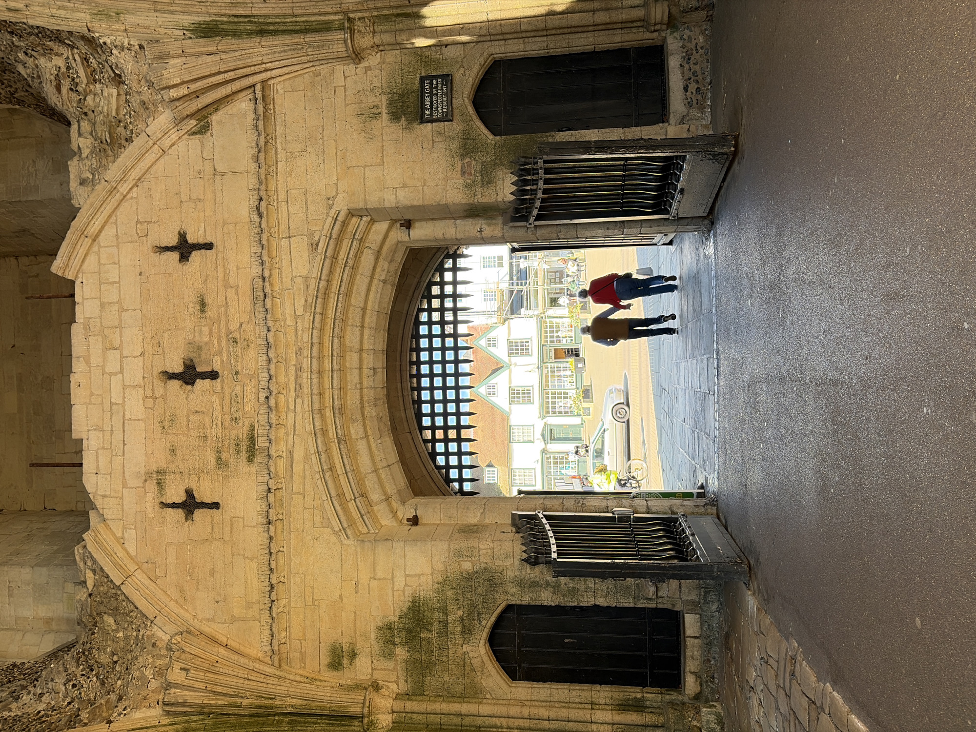 The image depicts the Abbey Gate in Bury St Edmunds, West Suffolk, United Kingdom. The gate is a large, medieval stone archway with a smaller arched doorway to the side. The stone is weathered and shows signs of age, including patches of moss. Iron gates are visible inside the archway. Two people are walking through the gate, one wearing a red jacket and the other wearing a yellow jacket. Outside the gate, buildings are visible, along with a parked car, scaffolding, and a person on a bicycle. The scene appears to be taken on a sunny day.