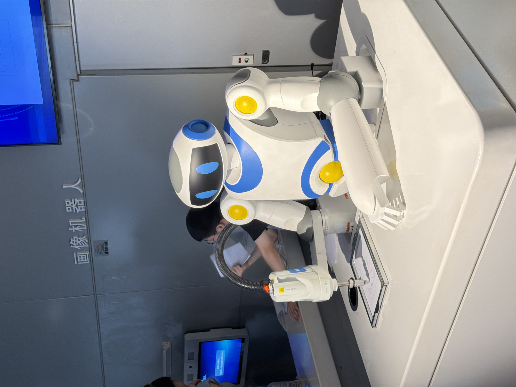 A humanoid robot with a white and blue body and yellow accents sits on a white surface. Its right arm is extended, holding a drawing tool that is sketching on a piece of paper placed in a clear frame. The robot is positioned in front of a gray wall with Chinese characters that read "图像机器人" (Image Robot). Below the characters is a small blue and white sticker. To the left of the robot, a white device with a flexible metal tube is mounted, also appearing to be part of a robotic system. In the background, two individuals are visible. One person, wearing glasses and a black shirt, is interacting with a monitor. The other person, partially visible, is standing near the monitor. A blue screen displays text. The environment appears to be an exhibition or demonstration space, likely located in 江北城街道, China, given the presence of Chinese text and a likely demonstration of advanced technology.