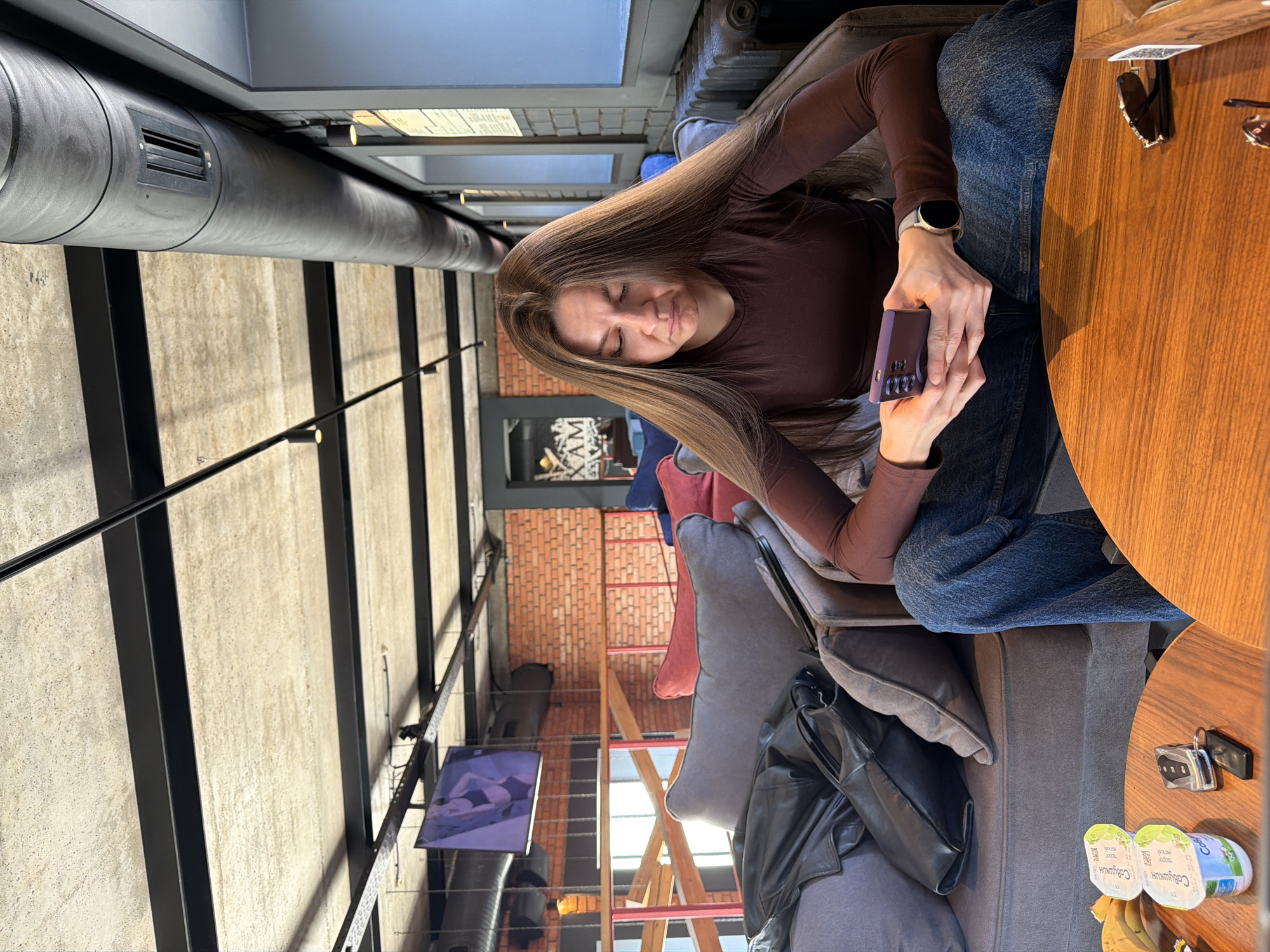 Here's a description of the image:
A woman with long, straight, light brown hair sits at a wooden table, holding a purple electronic device, likely a phone or a gaming console. She is wearing a brown long-sleeved shirt and blue jeans. She appears to be focused on the device.
The table is circular and made of wood, with a pair of sunglasses placed on it. Next to her, there is a grey couch with a black leather jacket and a grey pillow. On the table near her is also a set of keys and a yogurt cup.
The background reveals a modern interior with industrial elements, including exposed piping and concrete walls. A television screen is visible on the wall, showing a couple embracing. Red brick walls and scaffolding suggest the location is a cafe or lounge with an industrial-chic design.
Based on the image content, it is highly probable that the location is in a modern urban setting, specifically a cafe or lounge in Kazan, Russia, considering the cafe's design.
