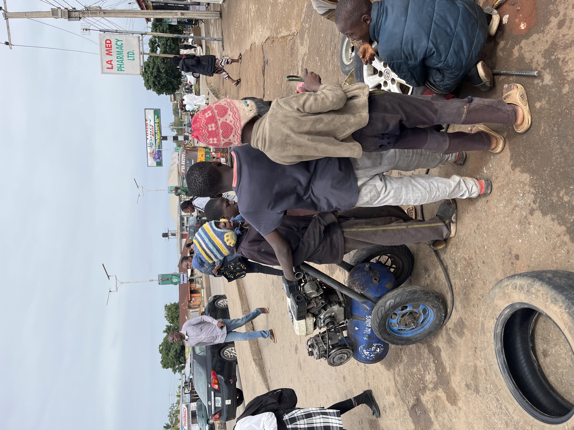 This street-level image captures a lively scene in Gura Top, Nigeria, featuring multiple individuals engaged in various activities. The setting is an urban or semi-urban environment with paved but worn ground, buildings, and overhead utility lines, under an overcast sky suggesting daytime.
The primary activity in the foreground involves tire repair. A person is crouched down, seemingly working on a car wheel that is detached from a vehicle. Nearby, a blue, engine-powered air compressor with "the blues" written on it and an old tire lie on the ground, confirming the service. Several men and a child in a red and white knitted hat stand around this central activity. Another child in a blue and yellow striped knitted hat is visible near the compressor.
Other people populate the scene: a man in a light shirt and jeans walks past a black sedan, and various pedestrians are scattered further down the street. Parked and moving vehicles indicate a busy thoroughfare.
Visible text on signs provides additional context: "LA MED PHARMACY LTD." on a white sign, "TRUST DANLADI ABOK ATU" on a billboard, and "Opay" and "Location Guard" on another sign. A small sign also displays "POS," and a vehicle has "PTA-11377" written on it. The overall atmosphere suggests a typical day of commerce and daily life.