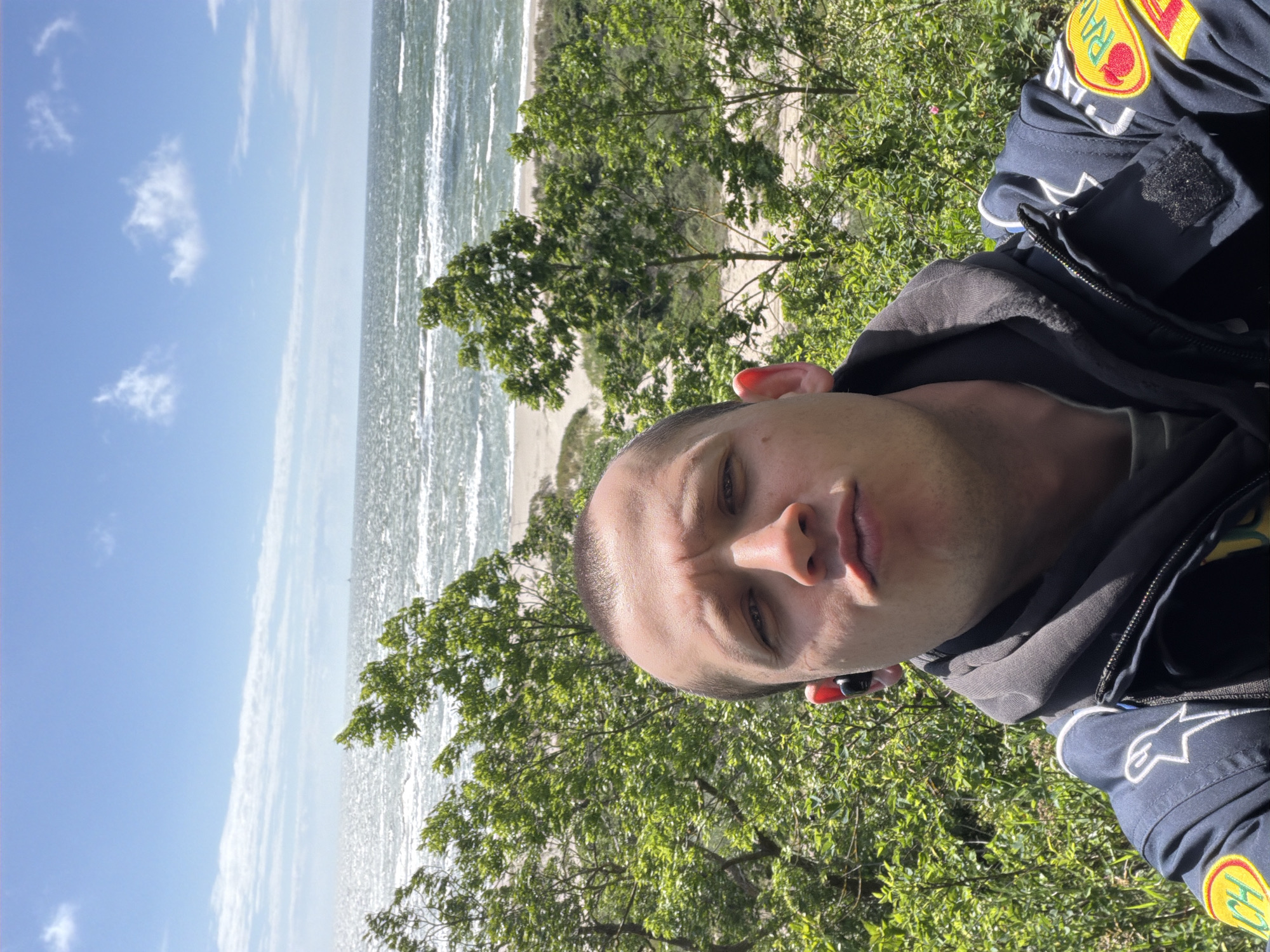 The image shows a person taking a selfie in front of a body of water with a beach and trees. The person is wearing a dark jacket with patches on the sleeve and appears to have an earbud in their ear. The sky is blue with some clouds. The location is identified as Povorovo, Russia.