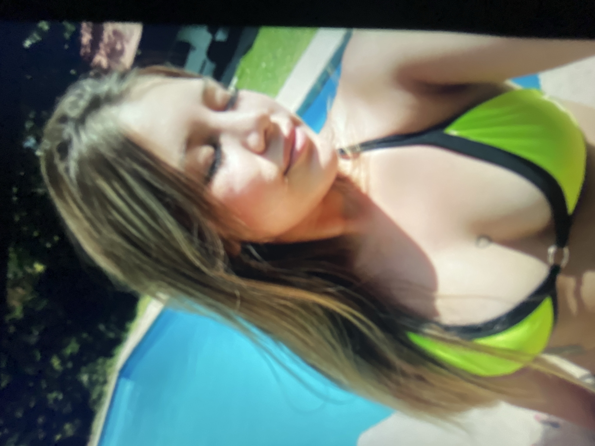 A young woman with long blonde hair is captured in a close-up selfie, posing by a bright blue swimming pool. She is wearing a neon green bikini with black trim. The background shows lush green foliage, suggesting an outdoor setting likely in a backyard or resort. Her eyes are closed, and she has a slight smile, giving the impression of relaxation and enjoyment on a sunny day. The scene is vibrant and captures a moment of leisure. This image does not provide enough information to determine a specific location within the city of Sofia, Bulgaria.