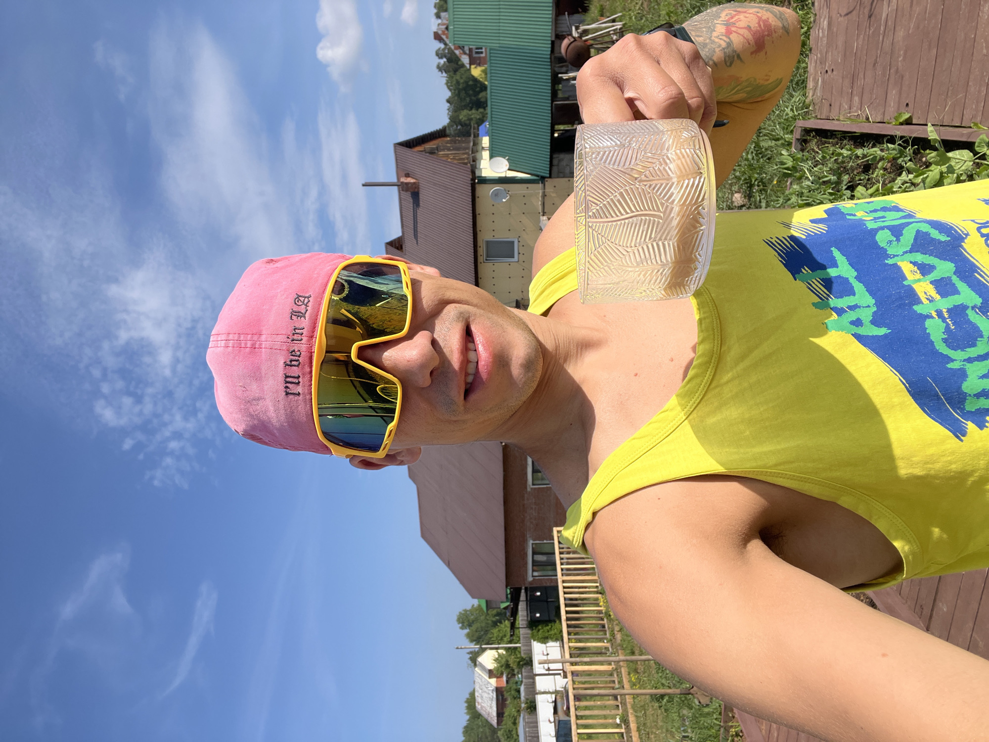 A man stands outdoors, holding a clear glass with a light-colored liquid. He is wearing a pink baseball cap with the text "I'll be in LA" embroidered on it, and bright yellow sports sunglasses. His shirt is a yellow tank top with a graphic design featuring the words "ALL INCLUSIV". The man has tattoos on his left arm. The background shows a residential area with several houses, some under construction, with wooden fences and greenery. The sky is blue with scattered clouds. The scene appears to be set in a rural or suburban area, potentially in the city of Chaikovsky, Russia, given the architectural style of the houses and the overall environment.