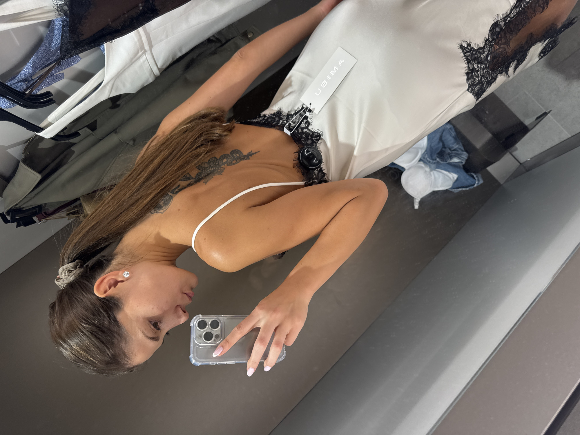 A woman is captured taking a mirror selfie from a high angle within a retail fitting room in Burgas, Bulgaria. She holds a smartphone, specifically an iPhone in a clear case, with her right hand. She is trying on a white satin slip dress accented with black lace trim along the bust and neckline. A product tag, displaying the brand "UZIMA," is attached to the dress. The woman has long brown hair, part of which is gathered with a scrunchie, and a tattoo is visible on her upper left back. She wears a diamond stud earring in her right ear, and her fingernails are manicured with light-colored polish.
In the background, to the left, a clothes rack holds several garments including a white top and a dark jacket. On the floor directly below the woman, a pair of jeans and another white clothing item are visible. The fitting room has neutral-toned walls and is illuminated by indoor lighting. The woman's expression is neutral and focused on taking the photograph.
