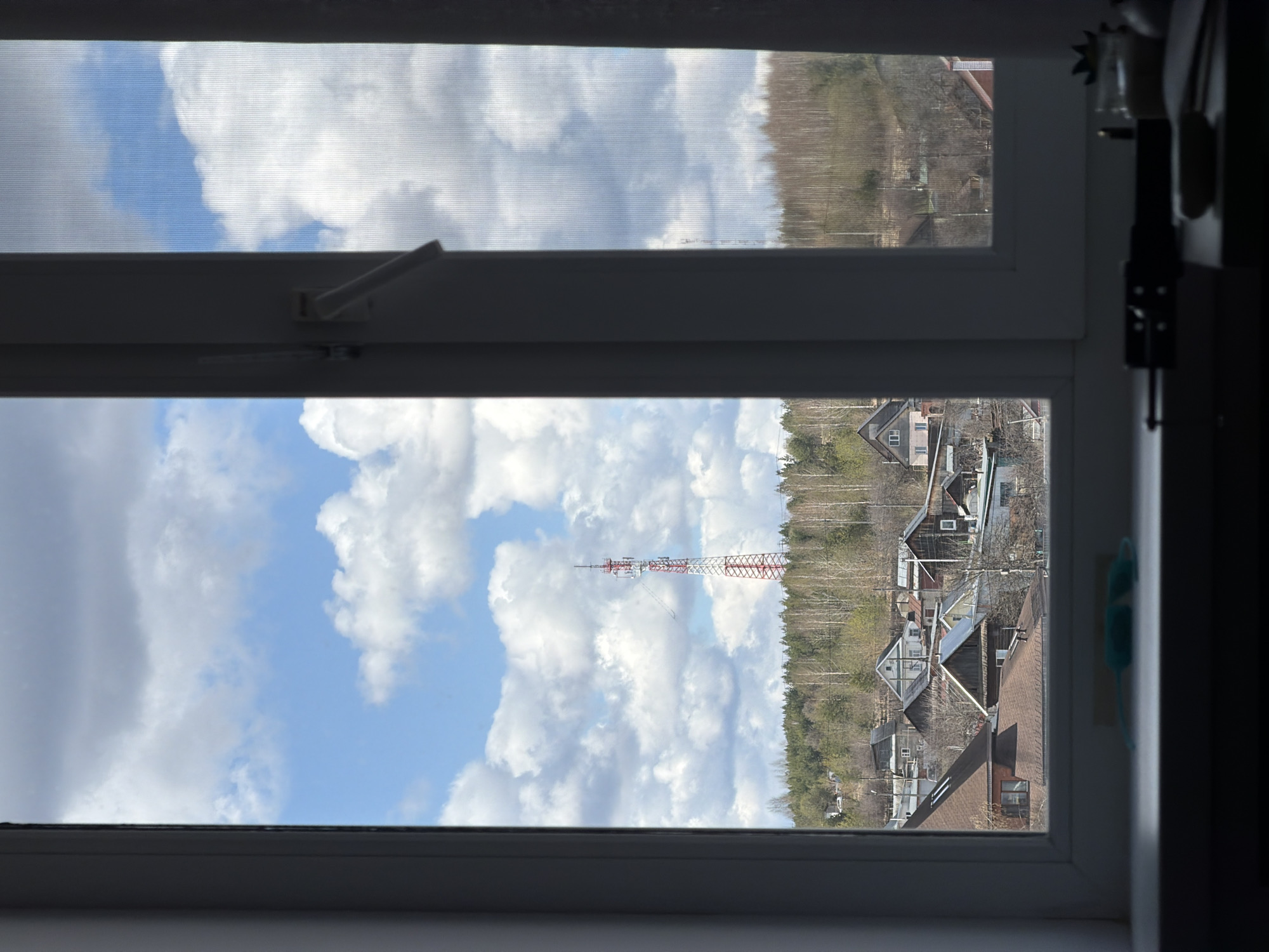 View from a window in Chaikovsky, Russia. The view includes a white window frame with a partially visible screen, a blue sky with white clouds, a red and white telecommunications tower, a forested hill, and houses.