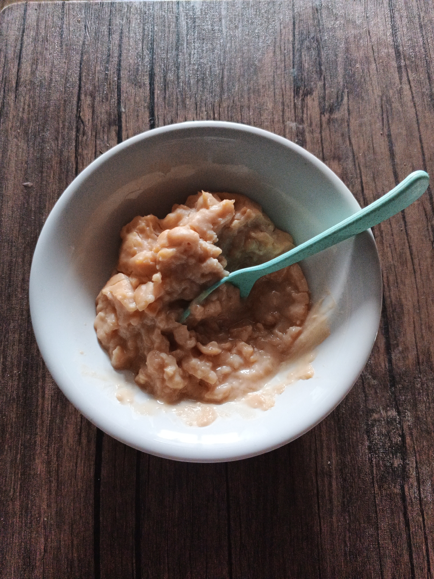 A light blue spoon rests in a white bowl filled with a thick, pale orange puree. The bowl is situated on a dark brown wooden surface with a visible grain pattern. The lighting suggests an indoor environment, possibly a kitchen or dining area. There are no people present in the image. The overall scene is simple and focused on the food. There is no information about the location in Gandasari, Indonesia.