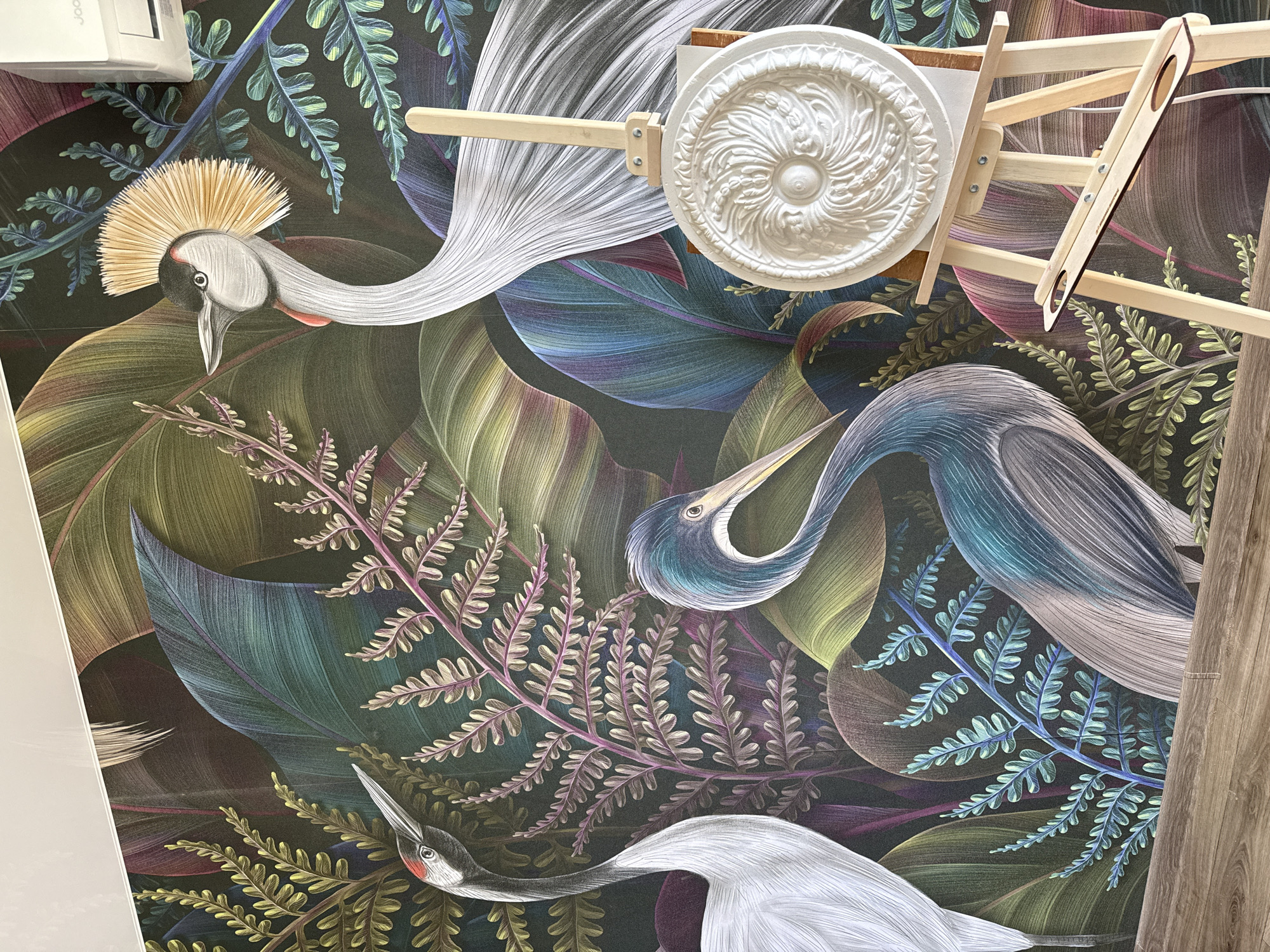 The image shows a close-up of a wall with a vibrant, detailed mural depicting a lush jungle scene with three stylized cranes and various tropical plants. The colors are rich and varied, with deep greens, purples, blues, and touches of yellow and red. In the foreground, a wooden easel stands with a white, ornate rosette ornament placed on it. The ornament has intricate, carved detailing. A wooden artist's tool with two circular cutouts rests on the easel's support. The background is the mural itself, which fills the entire wall. The lighting suggests an indoor setting, possibly a room or studio. The overall impression is one of artistry and decorative design. The location of Шолохово, Russia is not visually discernible from the image content itself.