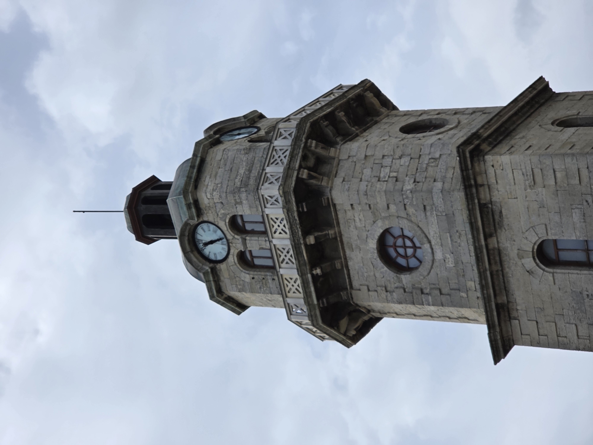 The image depicts the Clock Tower of Varna, Bulgaria, viewed from a low angle looking upwards. The tower is constructed of grey stone with a multi-tiered design, featuring clock faces on the upper sections. The clocks show the time, but the exact hour and minute is difficult to discern due to the angle. The tower is adorned with geometric decorative elements and has several arched windows. A thin metal rod protrudes from the top of the tower. The background is a pale sky with overcast clouds.
