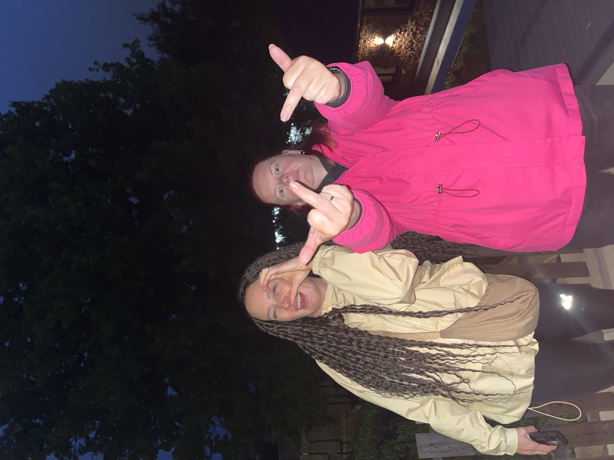 The image captures two adult females outdoors in the city of Chaikovsky, Russia, during low-light conditions consistent with evening or night. The female on the left has very long, dark, braided hair extending below her waist. She is wearing a light beige or off-white hooded jacket, dark leggings, and a light brown crossbody bag. Her right eye is closed in a wink, and her mouth is open in a wide smile. Her right hand is raised, with her thumb and index finger forming a circle and other fingers extended. She holds a dark-colored smartphone in her left hand. The female on the right has reddish hair and is wearing a bright fuchsia hooded jacket with adjustable drawstrings at the waist, dark undergarments visible at the neck, and dark pants. She has a visible nose piercing, a ring on her left ring finger, and a dark-colored watch on her left wrist. Both of her hands are raised towards the viewer, each displaying an extended middle finger. The background features dark trees against a dark blue sky. A wooden fence or railing is positioned behind the subjects. In the lower right portion of the background, a brick wall with two illuminated wall sconces is visible, alongside dark blue structural elements. The subjects are standing on a surface that appears to be a wooden deck or similar outdoor flooring.
