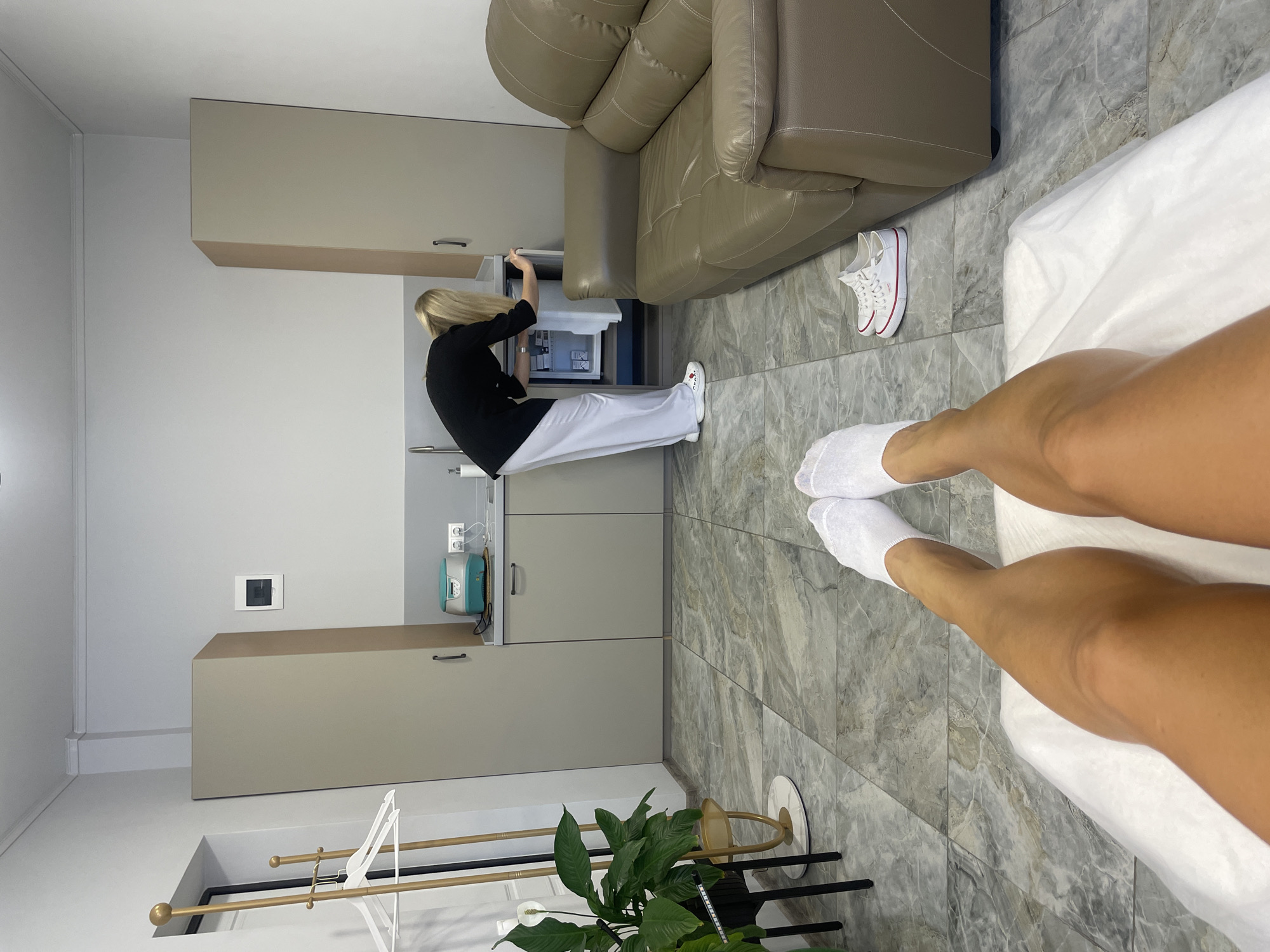 The image captures an indoor scene, likely a treatment room or clinic, from a first-person perspective looking across the viewer's legs. The viewer's tanned legs, clad in white socks, rest on a white sheet-covered surface, presumably an examination table or bed. The floor is covered with grey marble-patterned tiles.
In the background, a blonde-haired person, dressed in a black top and white pants, is seen from behind, bending over at a light beige counter with integrated cabinets and a sink. This person appears to be accessing a small white appliance, possibly a mini-fridge or cooler, located within the lower cabinetry. Above the counter, an upper cabinet matches the lower units. To the right of this person, a large, comfortable-looking beige leather armchair or sofa is visible, and next to it on the floor are a pair of white sneakers with red accents.
The room features white walls. To the left of the counter area, a wall-mounted square light switch or control panel is present. Further left, a floor-standing rack with gold and black bars holds an unseen item. A lush green plant in a dark pot, resembling a Peace Lily, adds a natural touch to the sterile environment. A blue and white device with a hose, possibly medical or aesthetic equipment, is visible near a power outlet on the wall by the counter. The overall setting is clean and modern, suggesting a professional establishment in Sochi, Russia.