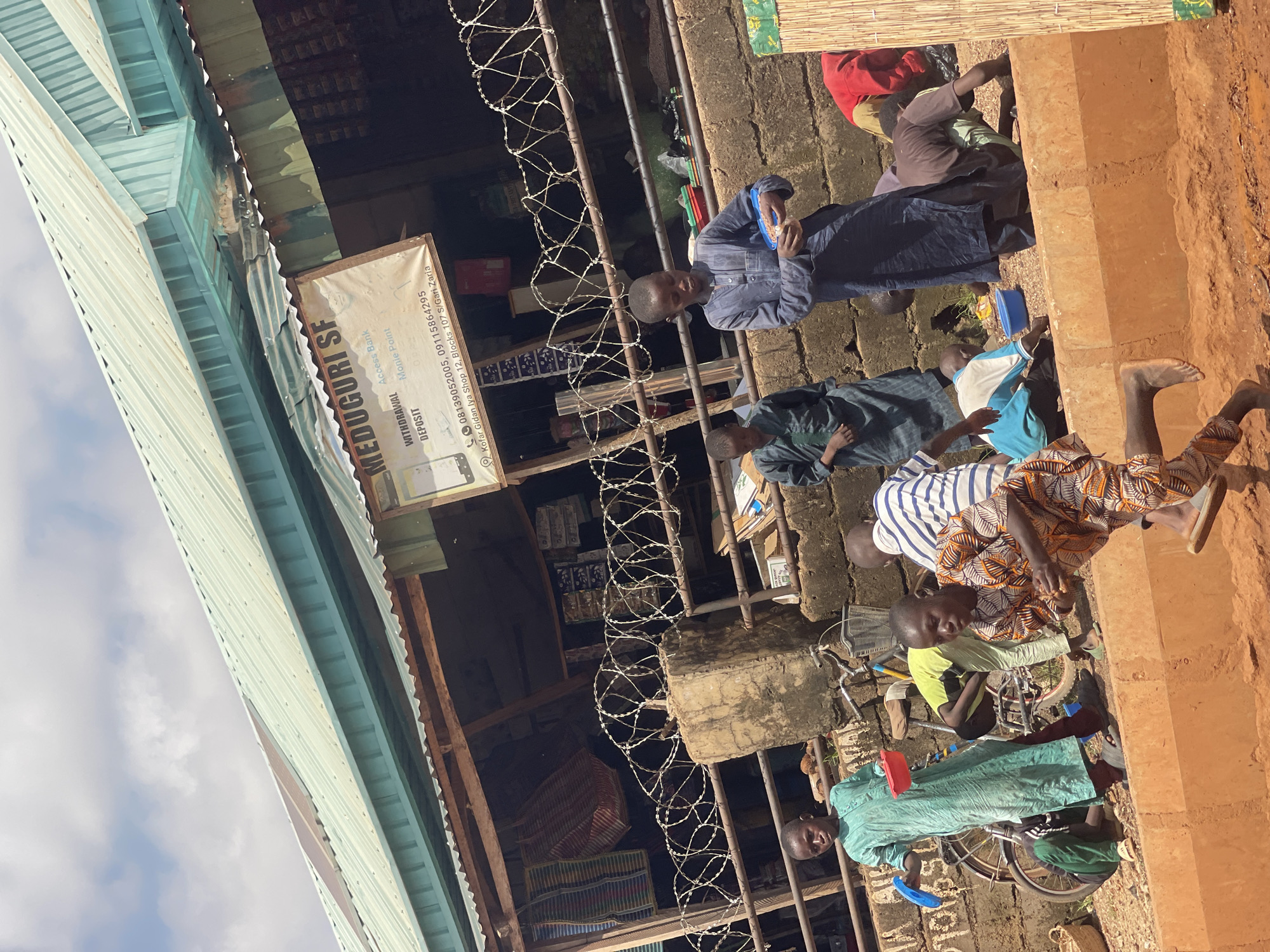 This is a medium shot of a group of children in an outdoor market setting in Zaria, Nigeria. The scene is dominated by a stall with a corrugated metal roof and wooden supports. A sign hangs from the stall reading "MEDUGURI SF" with additional text below indicating "WITHDRAWAL", "DEPOSIT", "Access Bank", and "Monie Point". Barbed wire is strung across the front of the stall.
Several children are present. In the foreground, a boy in a light blue, long-sleeved tunic is standing and looking towards the left. To his right, another boy in a dark blue, long-sleeved tunic is holding a small container and appears to be eating. Behind them, a boy in a striped shirt is partially visible. Further back, a boy in a light green tunic is holding a red bowl and a blue object. A bicycle with a basket is leaning against a brick wall behind the children. Other children are engaged in activities on the ground, including one in an orange and white patterned tunic bending over. A person wearing a red garment is also visible in the background, as is a stack of what appear to be boxes. The ground is dusty and uneven. The overall impression is of a bustling marketplace with children actively involved in their surroundings.