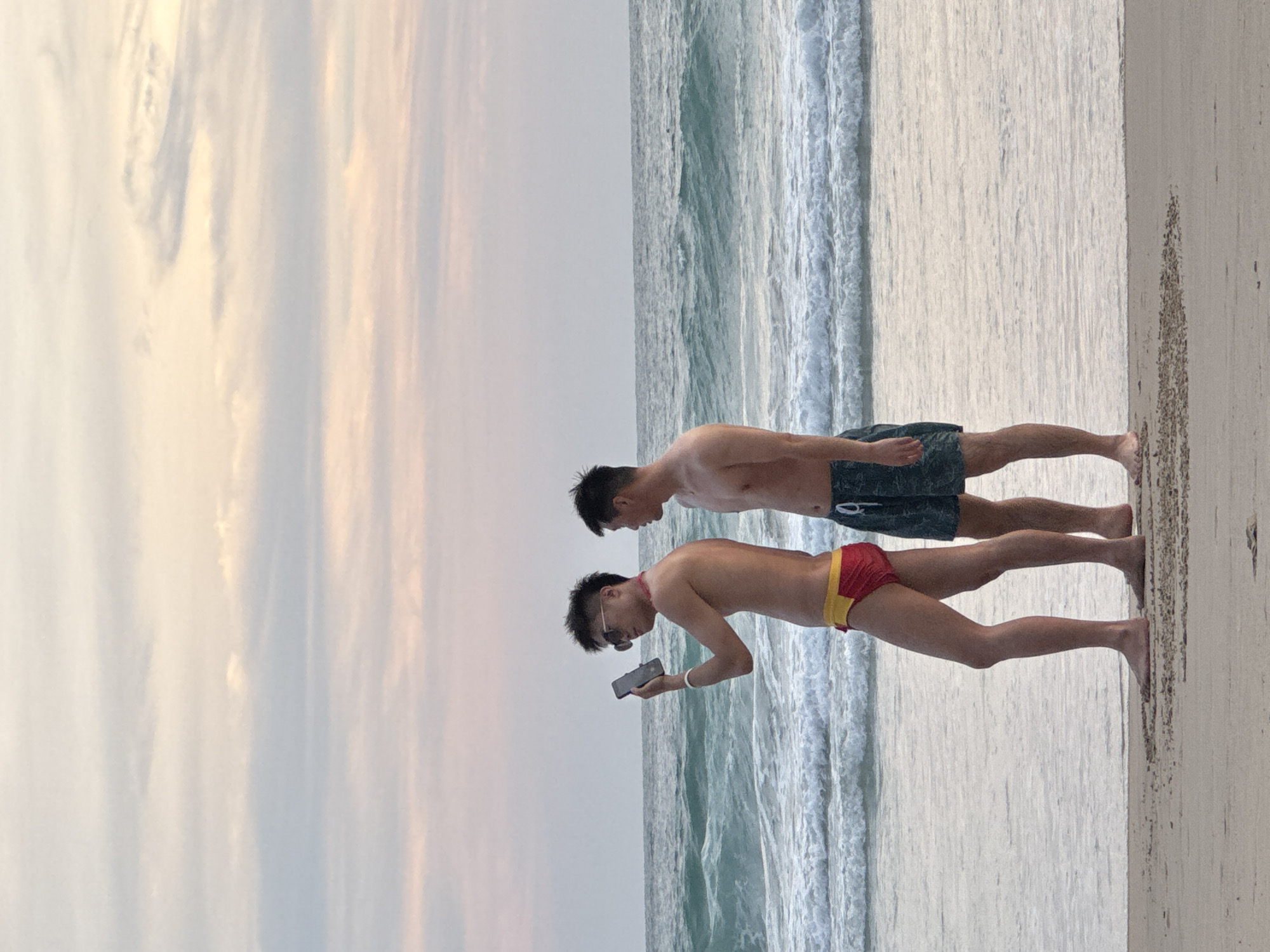Two bare-chested men stand on a sandy beach in Karon, Thailand, facing the ocean. The man on the left wears red and yellow swim trunks and sunglasses, holding a smartphone in his right hand. The man on the right wears dark patterned swim trunks. The ocean has moderate waves and the sky is overcast with pastel hues.