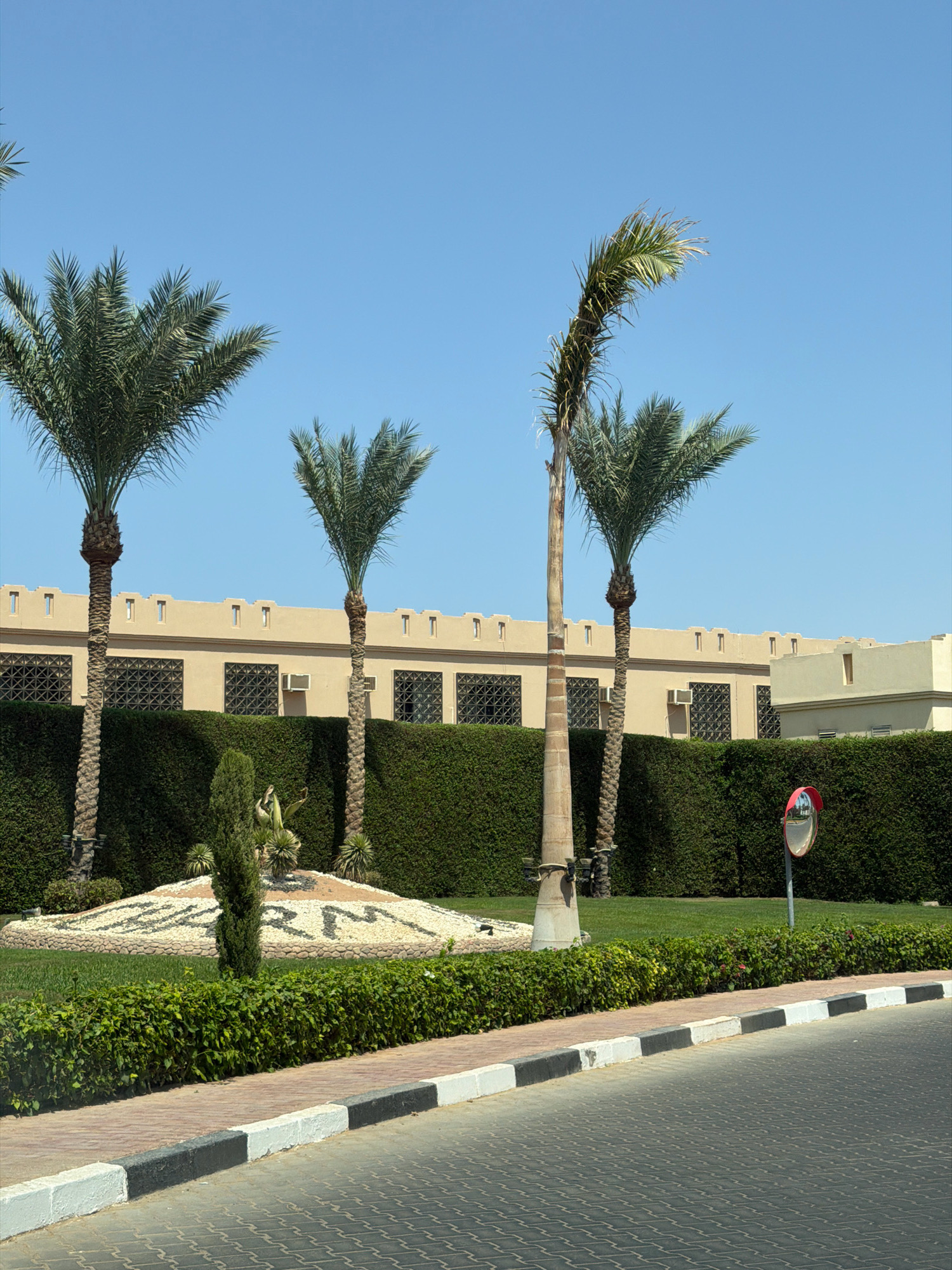 The image depicts an outdoor scene in Sharm El Sheikh, Egypt. In the foreground, a paved road with distinctive patterned grey bricks is visible, bordered by a curb painted with alternating black and white sections. Adjacent to the curb is a sidewalk constructed from reddish-brown pavers. Beyond the sidewalk, a neatly trimmed green hedge forms a barrier.
Further into the scene, a circular garden bed features a decorative arrangement of white stones and darker pebbles, forming the letters "SHARM". A slender cypress tree stands in the center of this garden bed, with other low-lying green plants surrounding its base. Two palm trees are prominent in the mid-ground, with one exhibiting a distinct curve and wind-swept fronds. Behind the hedge and garden, a beige building with a crenellated rooftop and dark-latticed windows runs horizontally across the frame. Several air conditioning units are mounted on the building's exterior. The background is dominated by a clear, bright blue sky. A convex security mirror on a pole is positioned to the right of the garden bed, reflecting the surrounding environment. The overall impression is of a well-maintained public space or resort area.