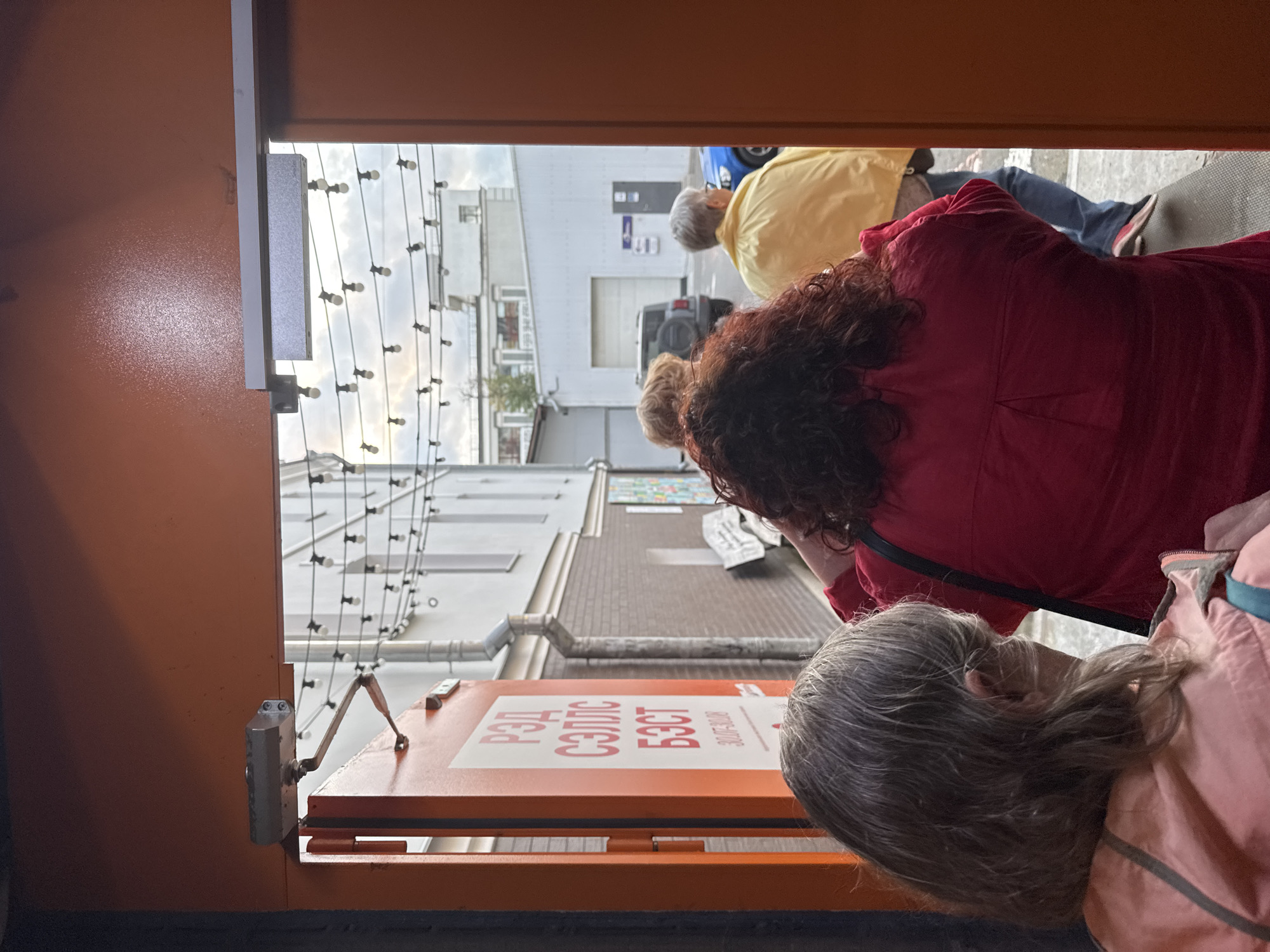 The image shows a rear view of three people standing in line to enter a building through an orange doorway. The person in the foreground on the left is wearing a pinkish-orange hooded jacket. The person in the middle is wearing a bright red top with curly dark hair. The person on the right is wearing a bright yellow jacket. Beyond the doorway, there is an alleyway with buildings, a grey SUV, and a blue car. String lights are visible hanging across the alleyway. A sign on the orange door has Russian text. The scene appears to be in Moscow, Russia.