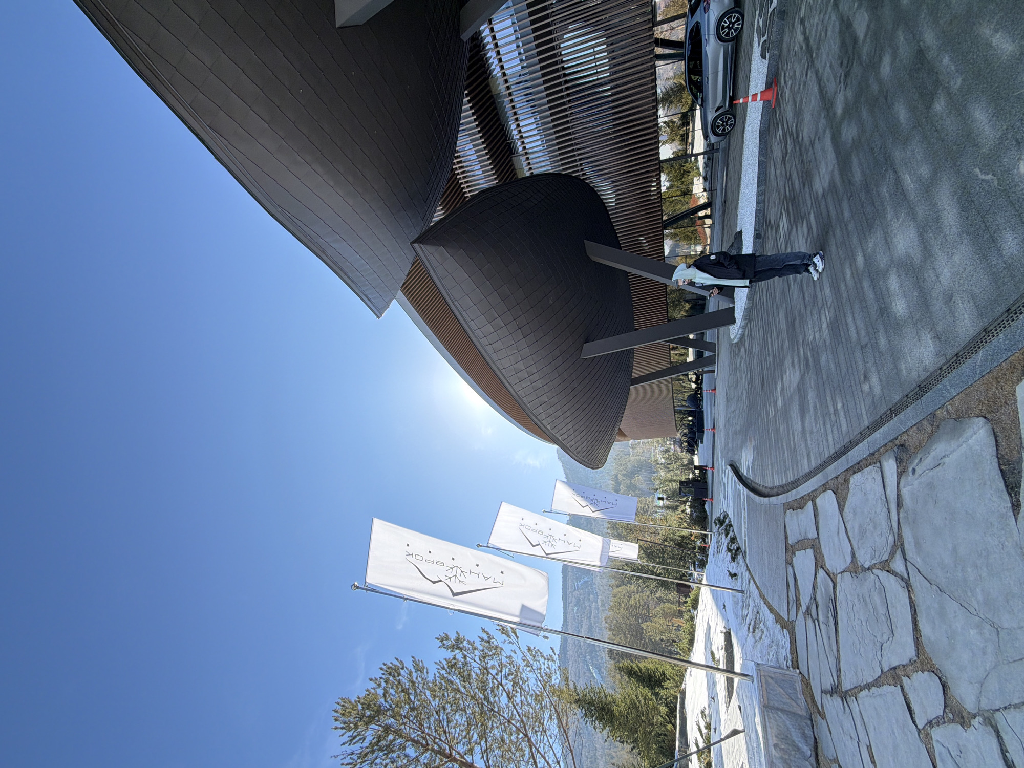 The image depicts the exterior of the Mayak Ecopark in Озёрное, Russia. A person stands on a paved walkway next to a gray car and an orange traffic cone. Above them, part of the unique, modern architectural design of the Mayak Ecopark structure is visible, featuring dark, layered roof sections and wooden slat paneling. Below, a series of white flags with a mountain logo and the words "МАЯК ЭКОРОК" in Russian are visible, their poles lining the pathway. The foreground shows rocky terrain interspersed with shrubs and greenery. In the background, a mountain range is visible under a clear blue sky.