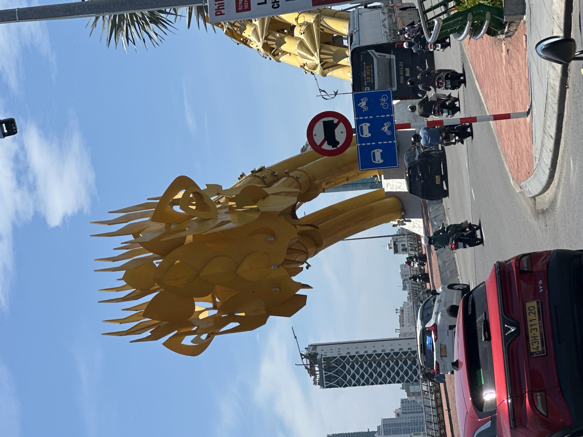This is a street-level, medium-angle, outdoor, daytime photograph taken in Da Nang, Vietnam, focusing on the Dragon Bridge. The primary subject is the golden dragon sculpture adorning the bridge, which dominates the upper left and right quadrants of the image, extending towards the center. The dragon's head, with its distinctive spikes and scale-like details, is prominently displayed.
In the foreground, a bright red VinFast VF6 electric vehicle is parked, its rear facing the viewer. The car has a Vietnamese license plate that reads "43H-311.20". To the left of the red car, a white SUV with a Vietnamese license plate "43A-033.90" is also visible.
The middle ground of the image shows traffic on the bridge. Several motorbikes are in motion. One motorbike with a rider wearing a green jacket is moving away from the viewer on the right side of the road. Further down the bridge, three motorbikes are traveling in the same direction. A black SUV is also present in the traffic. A large bus is partially visible on the right side, with "LE HUY" and Vietnamese text visible on its side.
Traffic signs are present, including a red and white circular sign with a truck icon crossed out, indicating no trucks. Below this is a blue rectangular sign with white icons depicting cars, motorbikes, and bicycles, indicating permitted traffic.
The environment is urban, with tall buildings visible in the background, including a distinctive skyscraper with a patterned facade. The sky is a clear blue with scattered white clouds, and palm fronds are visible on the right side of the frame, suggesting a tropical or subtropical climate. The road surface is asphalt, and a red and white striped pole stands on the right side of the road, near the curb.