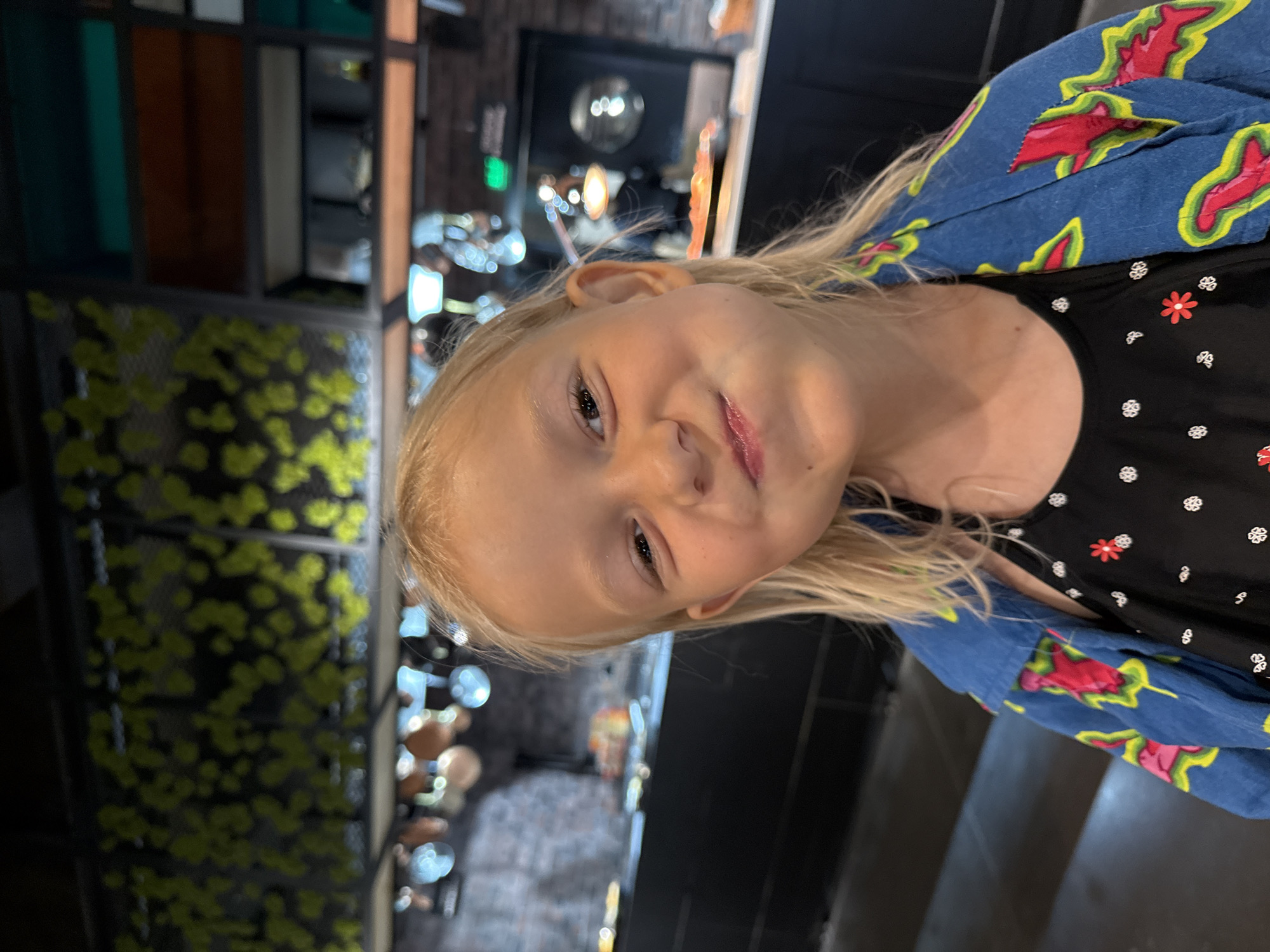 A young girl with blonde hair and fair skin is shown in a close-up, angled shot. She is wearing a black top with a pattern of small white and red flowers, and a blue jacket with a repeating pattern of stylized red and yellow flames or chili peppers. Her head is tilted, and she is looking towards the camera with a slightly playful expression, her lips slightly parted. Her eyes are open, and her gaze is direct.
The background is out of focus but appears to be an indoor setting, possibly a restaurant or a retail space. To the left of the girl, a section of a wall is visible, covered in a decorative pattern of small, bright yellow shapes that resemble scattered leaves or confetti. Behind her, in the distance, there are shelves with various objects and some blurred lights, suggesting a commercial or public environment. The lighting is ambient, and it's difficult to determine the time of day. There is no visible text in the image. The overall impression is a candid portrait of a child in a vibrant, perhaps slightly quirky, indoor environment.