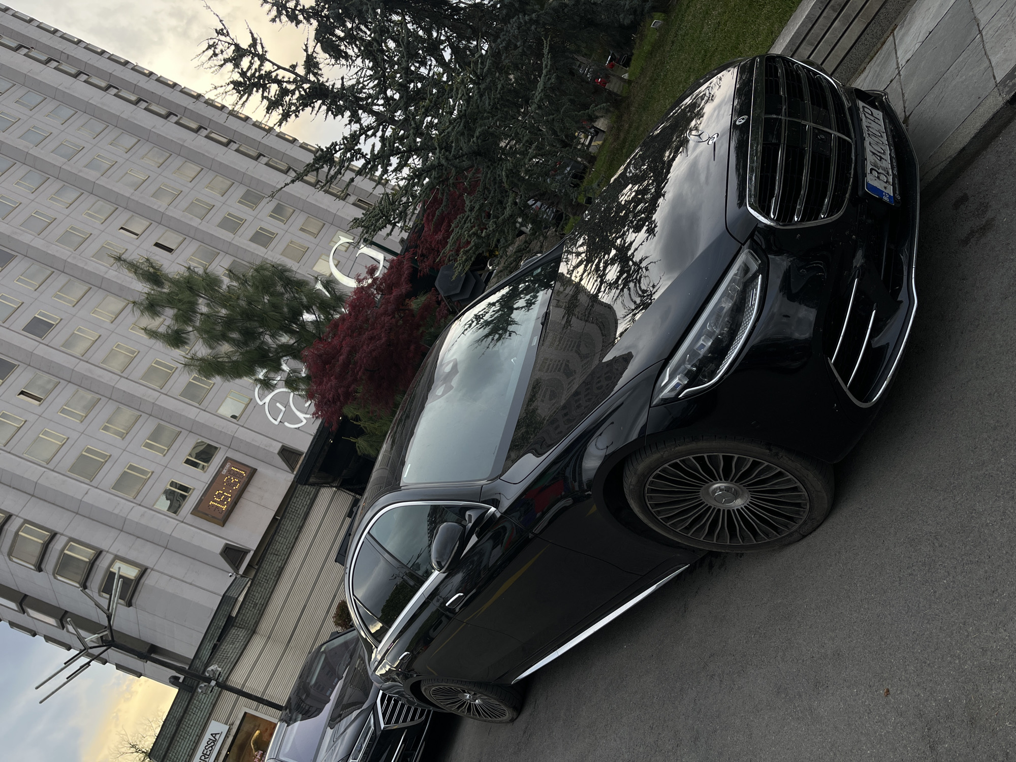 The image presents an urban street scene in Sofia, Bulgaria, captured during the mid-afternoon on an overcast day. A sleek, black Mercedes-Benz S-Class sedan (W223 generation) is prominently featured, parked on asphalt pavement. The car exhibits multi-spoke wheels, a large chrome grille with the Mercedes star on the hood, and modern LED headlights. Its front license plate displays "B 4000 TP," confirming the Bulgarian location.
In the background, a tall, light-colored building with a grid of numerous square windows rises towards the cloudy sky. A large, white letter "G" is visible as part of a sign on the building's facade. Below this, a digital display shows the time as "15:37." Further down, a partial shop sign reading "DRESSIA" can be seen. Various trees and bushes with both green and red foliage are interspersed throughout the frame, adding natural elements to the urban landscape. The sky is uniformly grey and overcast, its reflections visible on the car's polished surface and the building's windows. No individuals or specific activities are depicted, portraying a tranquil moment in a bustling city environment.