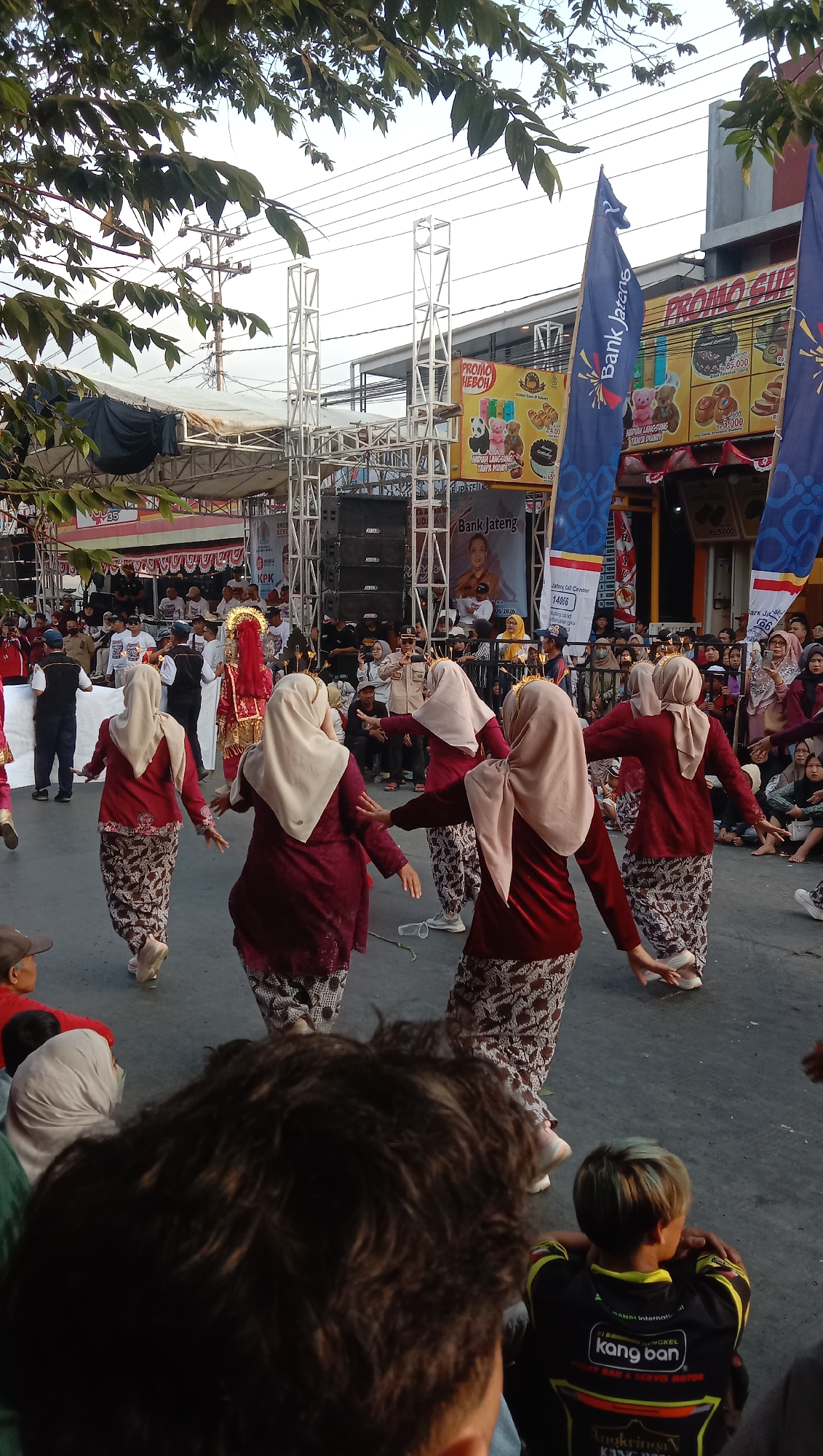 In Jatisawit, Indonesia, a group of women in maroon and cream hijab and patterned skirts perform a dance in a street festival setting. They are positioned on a grey asphalt surface, with a crowd of spectators gathered behind them. A stage with large speakers and a backdrop featuring the "Bank Jateng" logo and a portrait of a woman is visible in the background. Blue banners with the "Bank Jateng" logo are also prominently displayed. A person in a traditional red and gold costume, possibly a dancer or performer, stands slightly behind and to the left of the dancing women. The scene is outdoors, with trees and power lines visible overhead, suggesting an urban or semi-urban environment. The lighting indicates it is daytime.