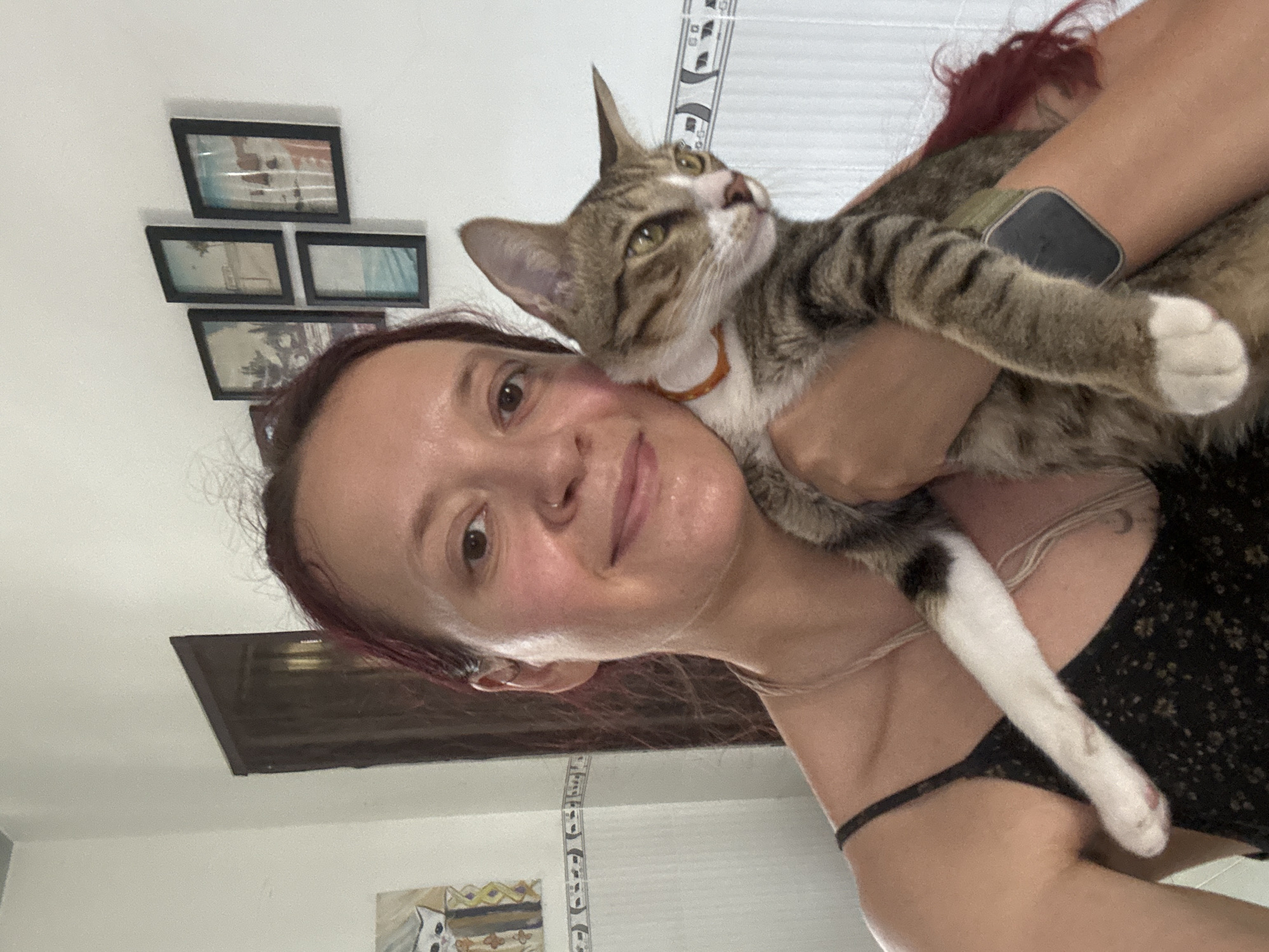 In Phu Thuy Ward, Vietnam, a woman with dark red hair tied back is holding a tabby cat. She is wearing a black spaghetti strap dress with a floral pattern. The cat has green eyes and is wearing an orange collar. The woman is smiling and looking at the camera. Behind them, on the wall, are four framed pictures of landscapes and a painting of a cat. A dark wooden door is visible to the left, and a tiled wall with a decorative border is on the right. The lighting appears to be natural, coming from a window or overhead source.