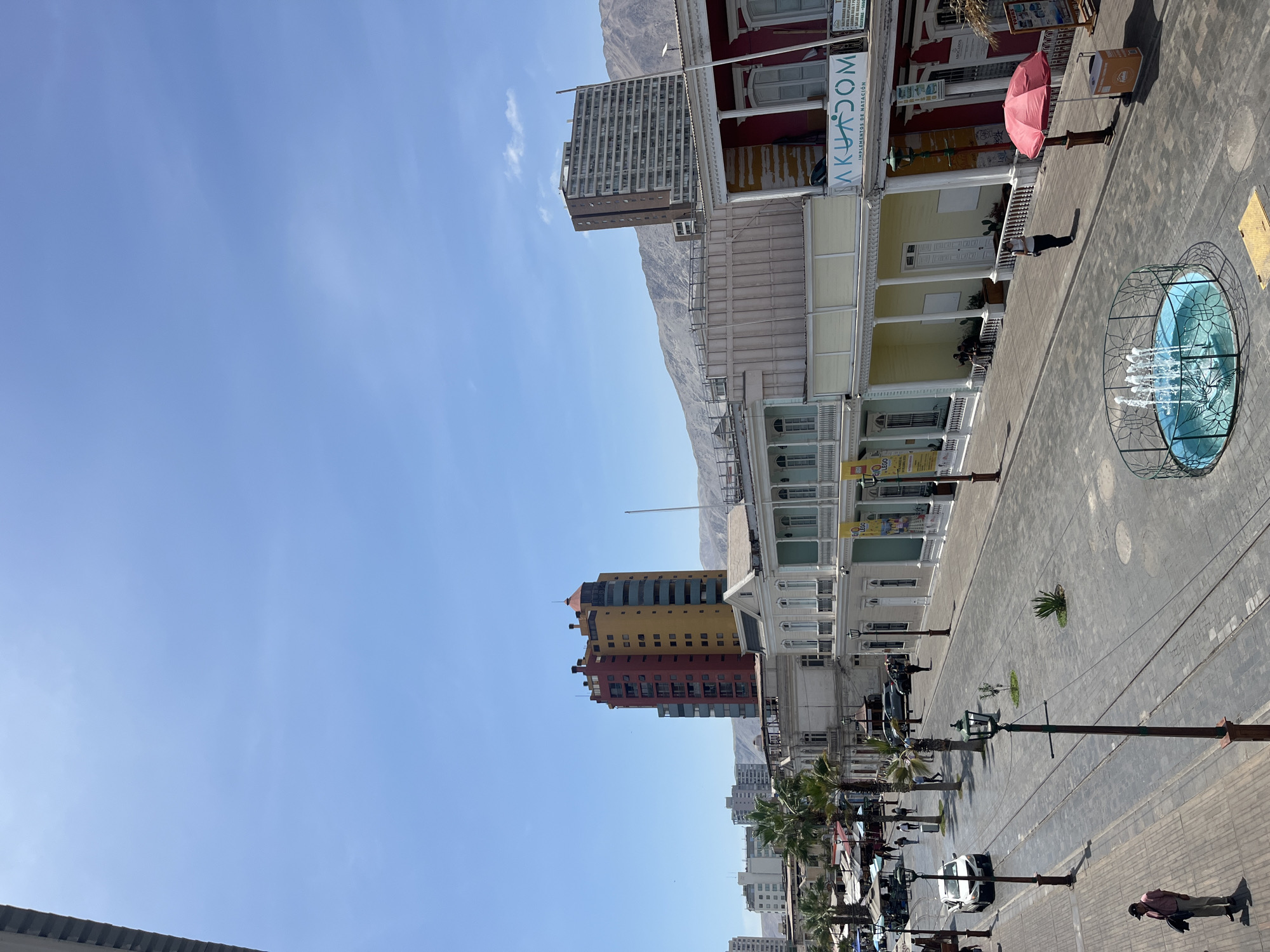 The image depicts a street scene in an urban environment. The location appears to be a coastal city with mountains visible in the background. Several buildings line the street, including a row of white buildings with colonial-style architecture, a tall modern building, and a building painted in shades of yellow and red. The street is paved with what seems to be cobblestones. A fountain is prominently featured in the foreground on the right. Street lights are placed regularly along the street. Cars and people can be seen moving along the street and sidewalks. A red parasol is placed next to a building.