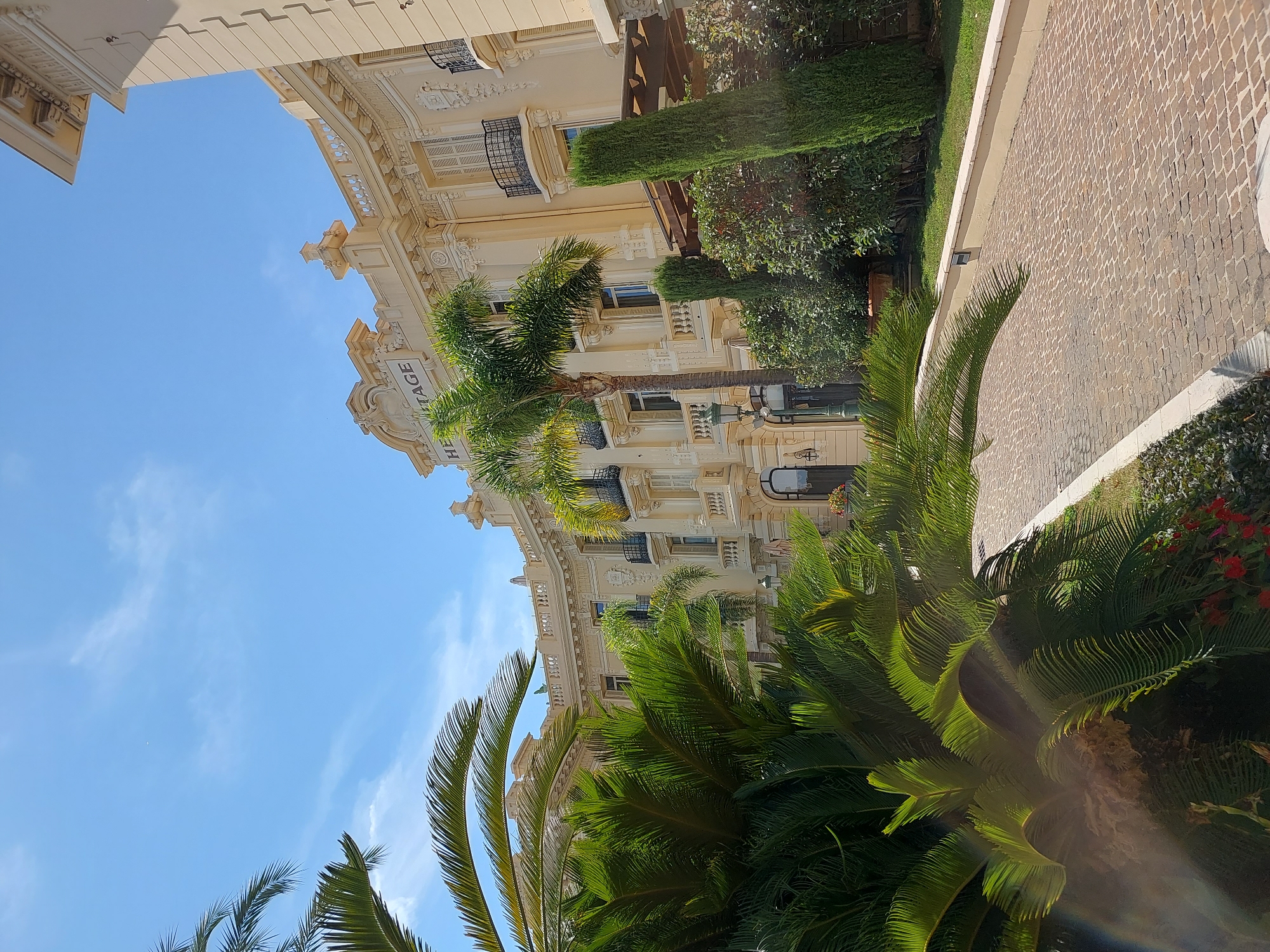 The image depicts the Hôtel Hermitage Monte-Carlo, a luxury hotel situated in the Carré d'Or district of Monaco-Ville, Monaco. The building features a light-colored, ornate facade with balconies and shuttered windows. In the foreground, lush green palm trees and other foliage create a tropical ambiance, with a cobblestone pathway winding through the scene. The sky is clear and blue with scattered white clouds, suggesting a sunny day. The hotel's name, "HERMITAGE," is visible on the facade. The composition, with its focus on the architecture and natural elements, conveys a sense of elegance and tranquility.
