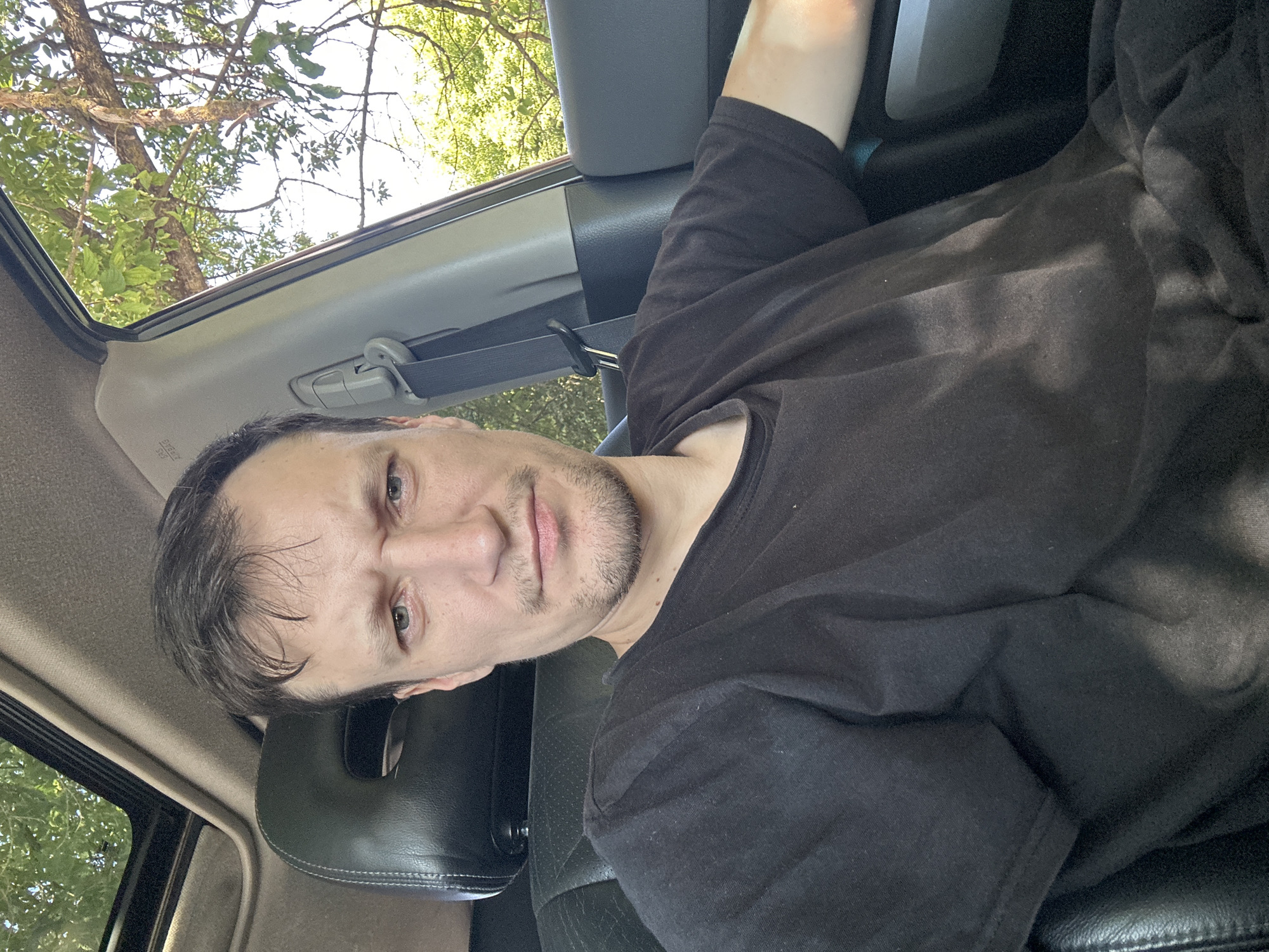 A man is seated inside a vehicle, his face captured in a selfie. He has short, dark hair and a light beard. He is wearing a black t-shirt. The interior of the car is visible, including a black leather seat, a sunroof, and a seatbelt. Outside the car, trees with green leaves are visible, suggesting an outdoor setting. The lighting indicates it is daytime, possibly sunny, with dappled light falling on the man and the car's interior. There is no specific indication of the location in Makiv, Ukraine.