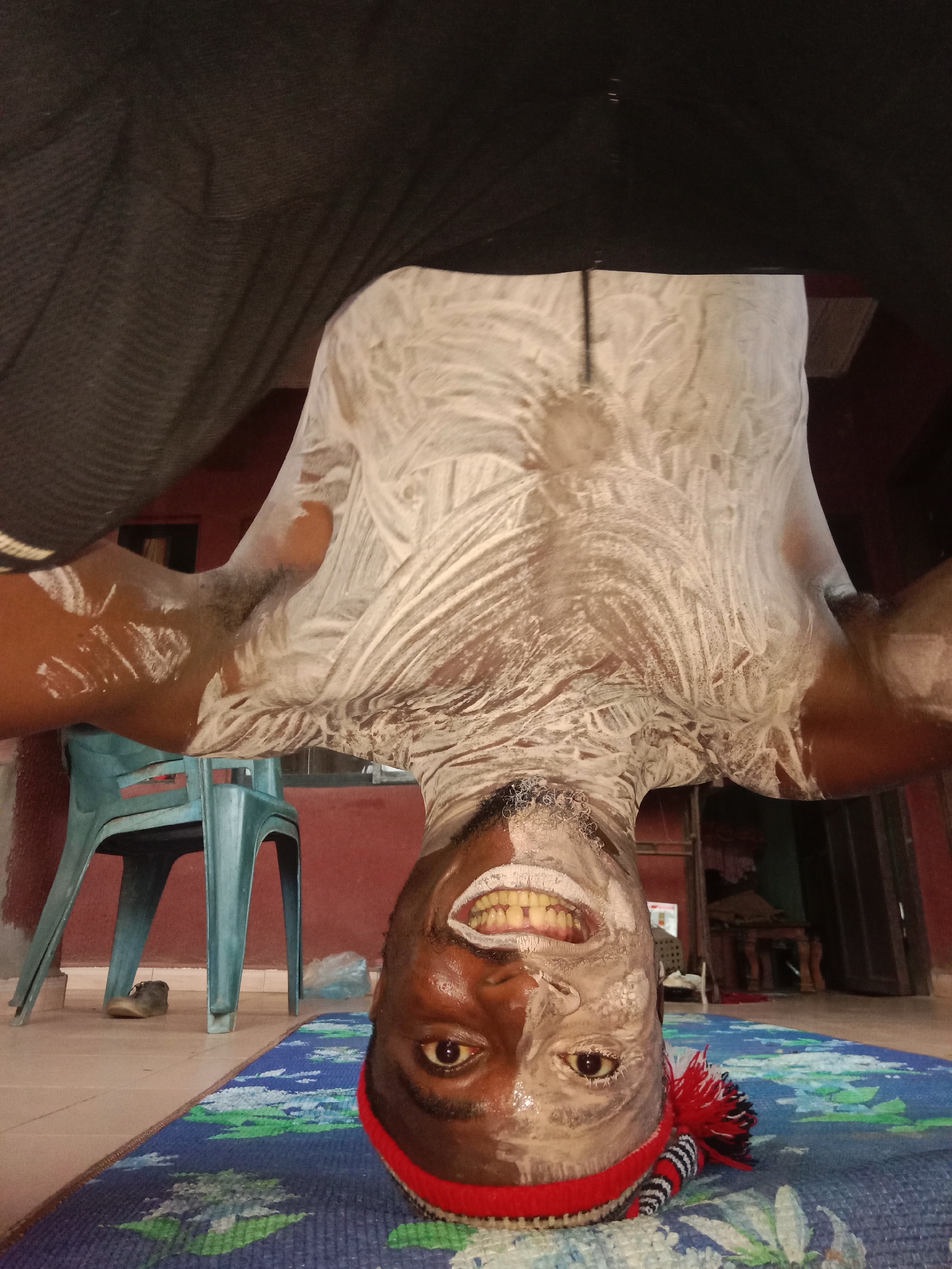 An upside-down medium shot reveals a Black man with a generous application of white substance on his torso, neck, and face. He is positioned on a mat with a blue and green floral design, and he wears a red and black striped headband with a red tassel. His eyes are open, and he is smiling, exposing his teeth.
Behind him, a blue plastic chair sits next to a red wall. A single brown shoe rests on the tiled floor. The background includes a doorway and a partial view of furniture inside, suggesting a domestic setting. The location appears to be indoors.