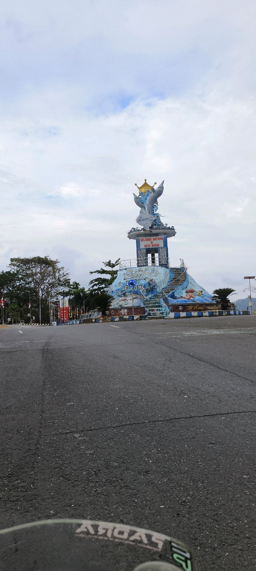 The image depicts a prominent monument in Waisai, Indonesia, a city known for its dolphin statues and as the capital of Raja Ampat Regency. The monument features two dolphins crowned with a golden crown, emerging from a sculpted base that resembles a rocky outcrop. The base is decorated with blue and white wave patterns and has the word "WELCOME" inscribed on it. Text on the monument reads "SELAMAT DATANG DI KOTA WAISAI" (Welcome to Waisai City) and "KABUPATEN RAJA AMPAT" (Raja Ampat Regency).
The monument is situated on the side of a wide, empty asphalt road, suggesting it's a roundabout or a roadside attraction. The surrounding environment includes trees, some palm trees, and flags. The sky is overcast with scattered clouds. The foreground shows the asphalt road with faint lane markings. The bottom of the image shows a reflection or part of a vehicle, possibly a motorcycle, with a logo that reads "YAGUAR" mirrored. The overall scene suggests a quiet, possibly early morning or late afternoon atmosphere.