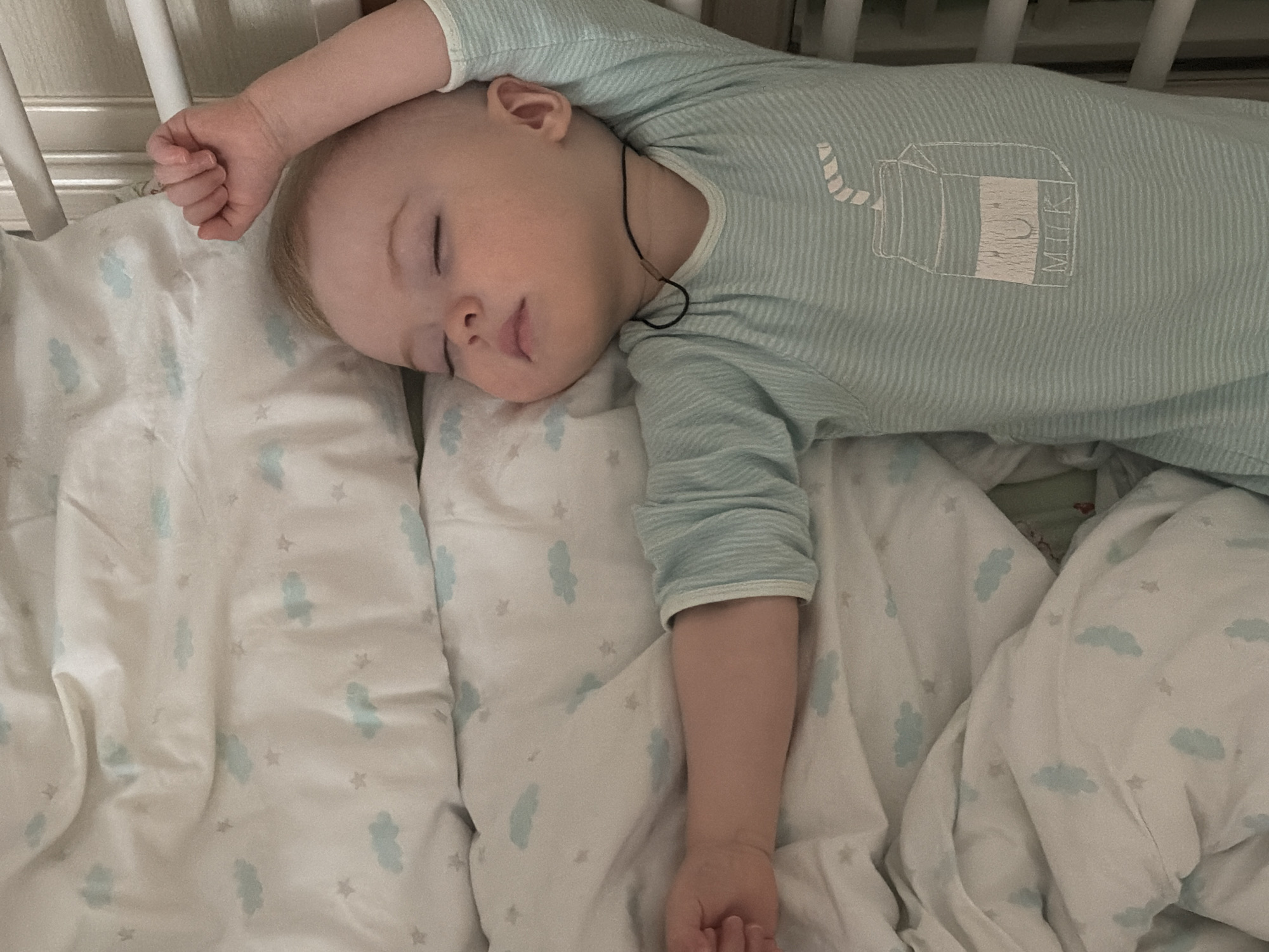 A baby wearing a striped light blue onesie with a milk carton design sleeps in a crib. The crib has white bars. The baby is lying on a white blanket with a pattern of blue clouds and small stars. A thin black cord is around the baby's neck. The location is unknown and there is no indication that this image was taken in the city of Votkinsk, Russia.