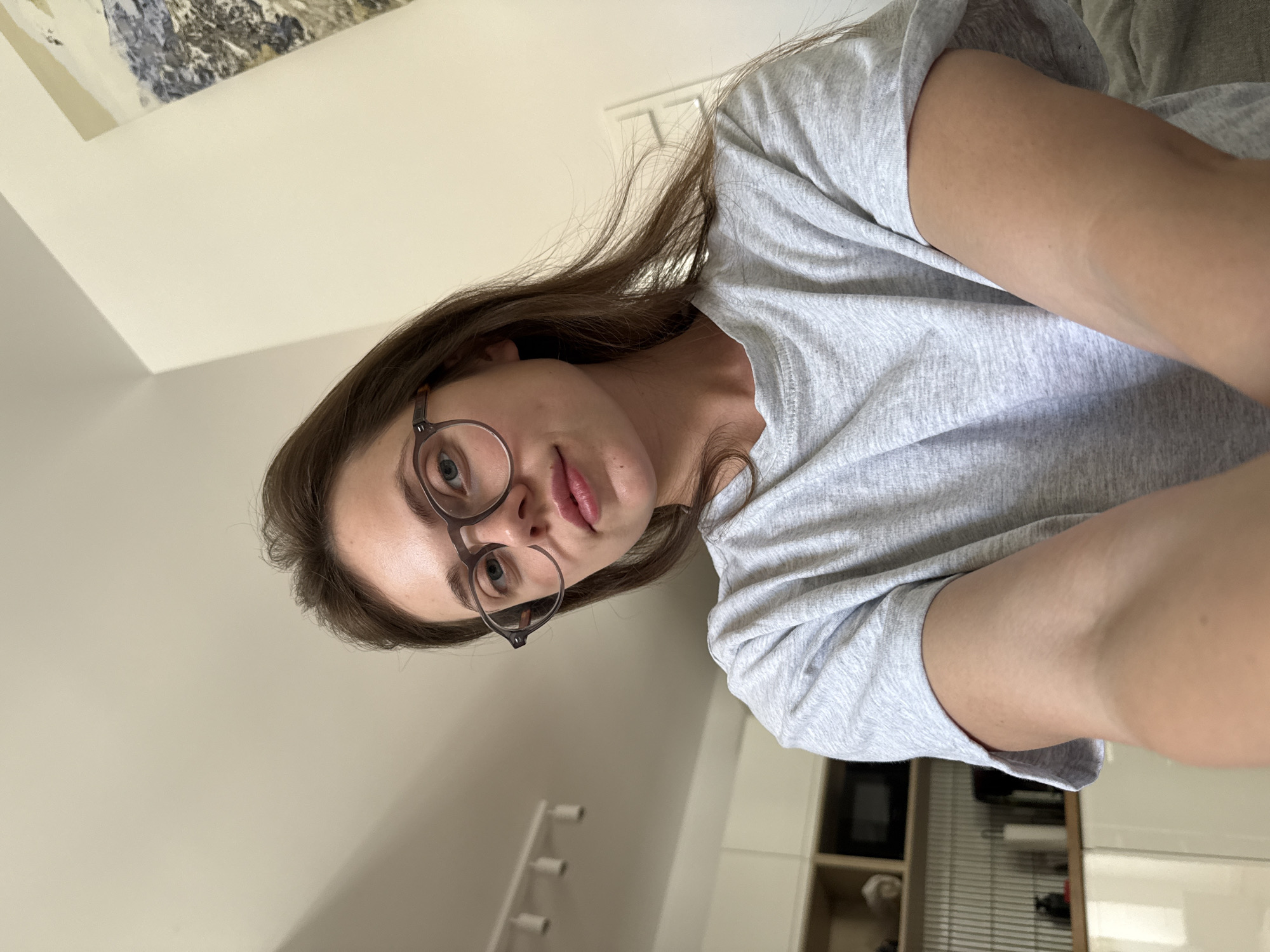 This is a vertical, medium-close-up selfie of a young woman wearing a grey t-shirt and glasses. She has long, brown hair and is looking directly at the camera with a neutral expression. The background appears to be an indoor setting, possibly a home, with a light-colored wall and a piece of abstract art on the wall in the upper left corner. The lighting suggests it could be daytime. There is no visible text in the image. The image is rotated 90 degrees clockwise.