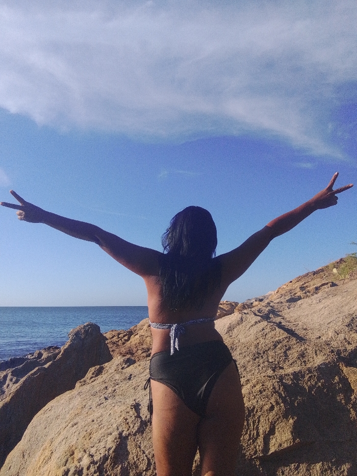 A woman stands on a rocky outcrop overlooking the ocean, her back to the viewer. She's wearing a dark bikini bottom and a light-colored top with ties. Her arms are outstretched, forming a "peace sign" with both hands. The sky is clear blue with a few scattered clouds. The lighting suggests it is daytime. The scene conveys a sense of freedom and connection with nature.