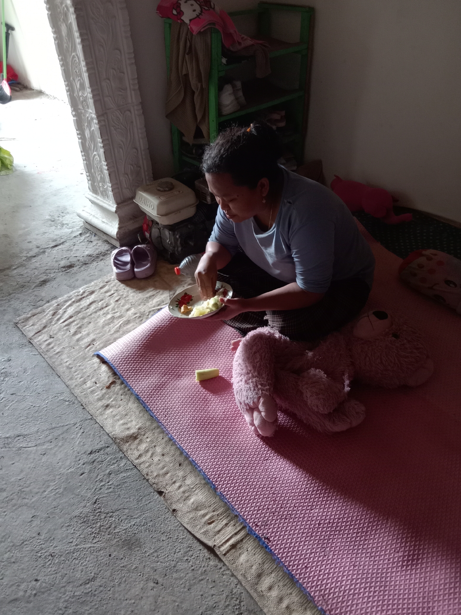 A woman is eating from a plate of food while sitting on a pink mat on the floor of a room. A large, light pink teddy bear is lying next to her. The room has a concrete floor and a white pillar on the left. There are also some shelves with items on them and a bottle of water. The woman is wearing a blue t-shirt and dark pants. The scene appears to be in a domestic setting in Pacing, Indonesia.