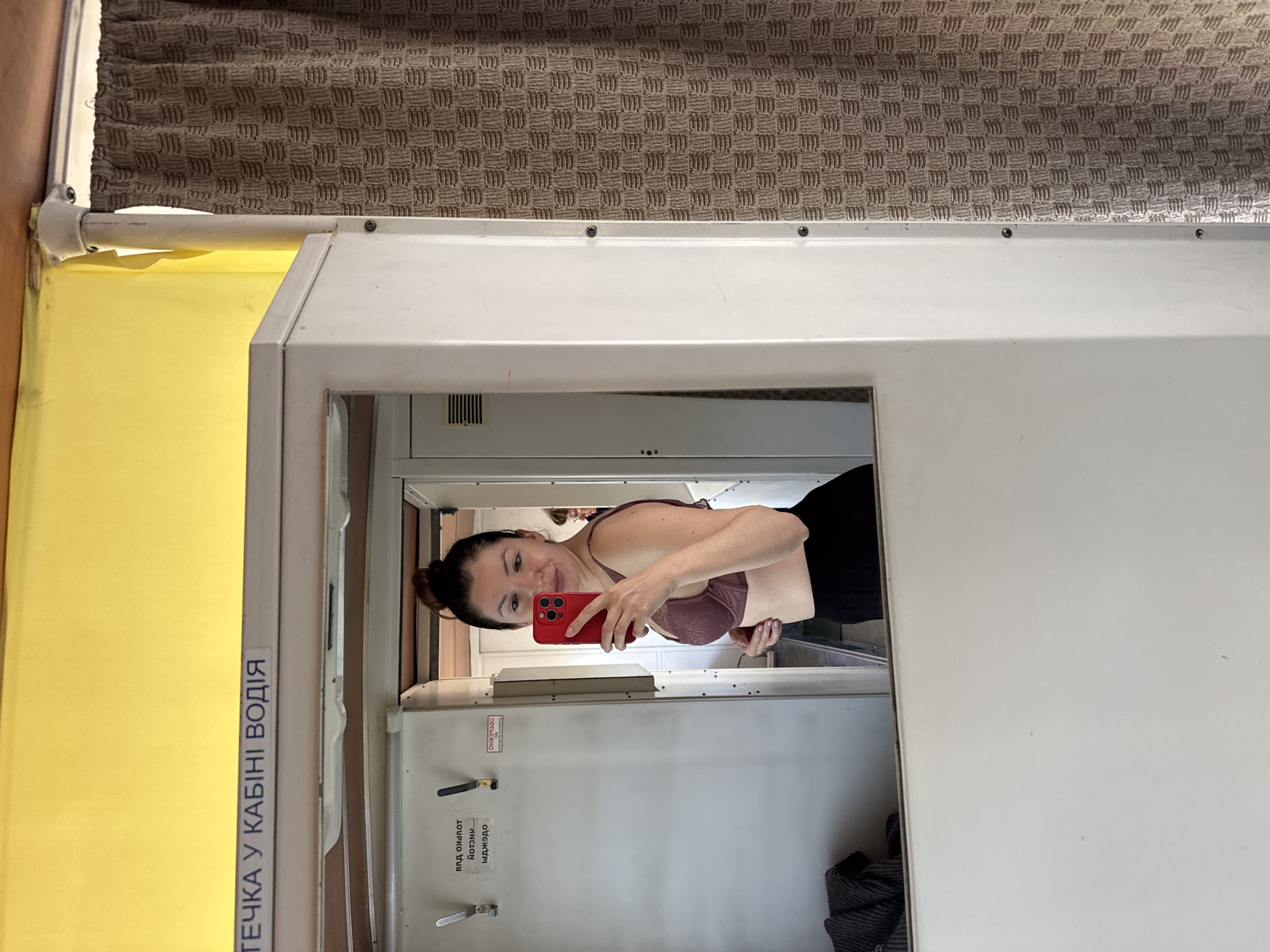 The image captures a vertical selfie taken by a woman in a mirror, revealing a functional indoor setting, likely a driver's cabin within a vehicle. The woman is positioned in the mirror reflection, looking towards the camera with a slight smile. She has dark hair styled in a bun and is wearing a dark pink or mauve bra and dark trousers or a skirt. She holds a red smartphone with multiple camera lenses to take the picture.
The mirror is set into a white, cabinet-like structure. Above this structure, a brown, textured fabric curtain or blind is visible. To the left of the white structure, there is a bright yellow panel or wall. On this yellow panel, a white label bears Cyrillic text: "ТЕЧКА У КАБІНІ ВОДІЯ," which translates to "FOLDER IN DRIVER'S CABIN." This detail strongly indicates the location is the driver's compartment of a vehicle, consistent with the user-provided context of Kryvyi Rih, Ukraine, a city known for its extensive public transport systems like trams and metrotram.
Reflected behind the woman in the mirror is a white door or panel with handles. Some reversed and partially obscured Cyrillic text is visible on this door, including "ЯКА ОНРОТ" and "ЯДЖАОТ," possibly indicating instructions or warnings related to the cabin or its operations. A small ventilation grill is also visible on one of the white panels. The overall lighting is artificial and bright, suggesting an indoor setting with no indication of the time of day. The interior design appears utilitarian and sturdy, typical of public transport vehicles. A dark, striped fabric item is partially seen at the bottom right of the mirror's reflection.