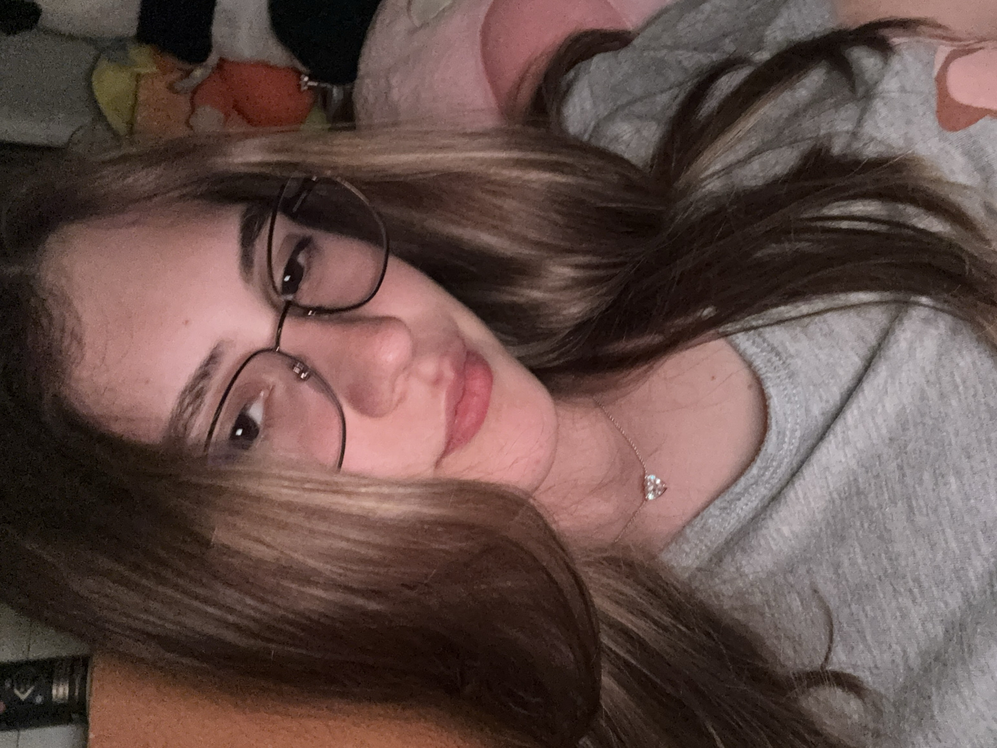 The image captures a young person, likely female, with long, wavy brown hair featuring lighter highlights, lying on their side and looking towards the camera. They are wearing dark-framed, round glasses and a light gray, possibly heathered, long-sleeved shirt. Around their neck is a delicate silver-colored chain necklace with a small, sparkling heart-shaped pendant. The person has a calm and relaxed expression, with a subtle, gentle smile on their lips.
The setting appears to be indoors, potentially a bedroom or living area in Panagyurishte, Bulgaria. The background shows soft textures and colors, including hints of pink and darker shades, suggesting pillows or blankets. A warm orange-brown surface is visible in the lower-left corner, possibly another blanket or bedspread. The lighting is soft and ambient, characteristic of an indoor environment. No specific activity beyond resting or relaxing is depicted. There is no visible text in the image.