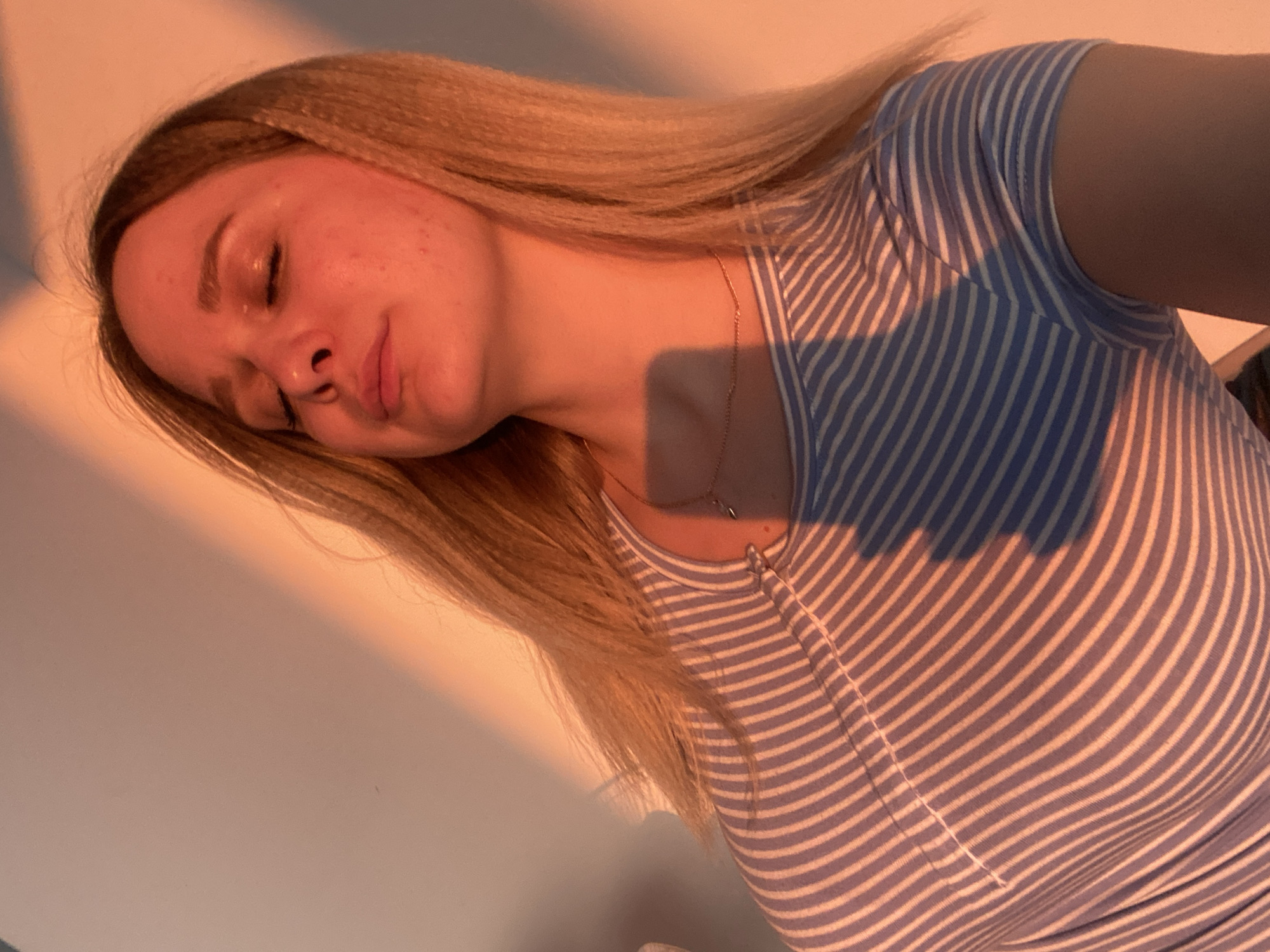 This media file features a close-up, tilted shot of a young Caucasian woman with blonde hair. She appears to be lying down or leaning, with her eyes closed and a relaxed expression. The warm, golden light suggests it is either sunrise or sunset. The woman is wearing a light blue and white striped t-shirt. A delicate necklace with a small, dark pendant is visible around her neck. The background is a plain, light-colored wall, with a prominent diagonal shadow cast across it and the subject, indicative of the low angle of the sun. The overall mood is peaceful and serene. No visible text is present in the image. The context provided places this scene in Izhevsk, Russia.
