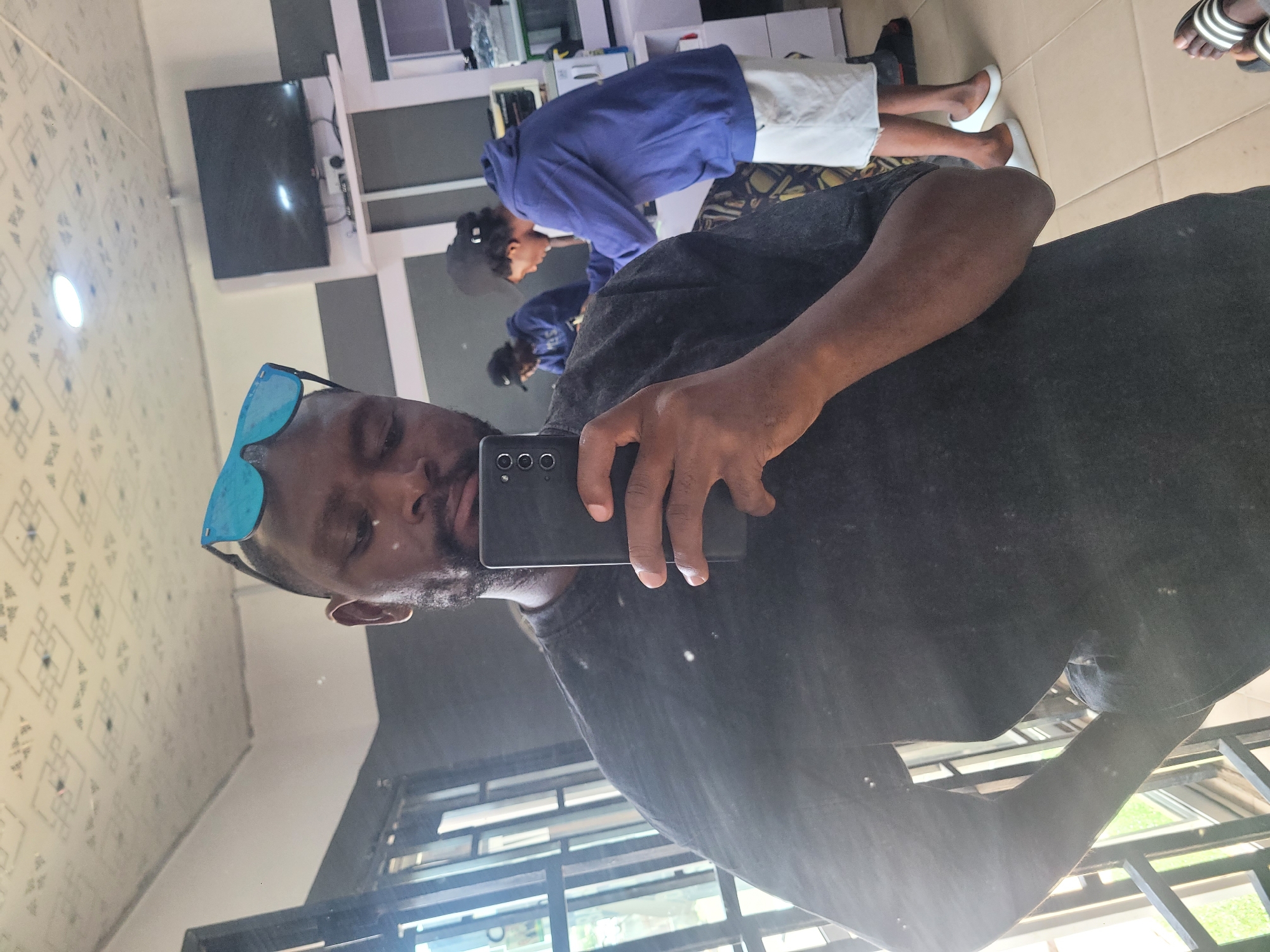 The image is a mirror selfie featuring a dark-skinned individual from the chest up, wearing a dark t-shirt. Blue mirrored sunglasses are pushed up onto their forehead, and they have a short beard or stubble. The person holds a black smartphone with their right hand, revealing its three-lens camera array, suggesting it's the device used for the selfie.
Reflected in the background, an indoor scene unfolds. Another person is visible, wearing a blue hooded sweatshirt and white shorts. This individual has dark hair styled back and possibly a cap, appearing to be standing or engaged in an activity further back in the room. The feet of a third person, wearing black and white striped slide sandals, are also reflected.
The setting features a light-colored, patterned floor and a wall that is partly light-colored and partly dark grey. A flat-screen television is mounted on a white shelf or wall unit, with what seems to be a media box placed below it. The room is brightly lit, indicative of daytime. The overall environment suggests a modern indoor space, potentially a living room, waiting area, or a salon in Jos, Nigeria. No discernible text is visible in the image.