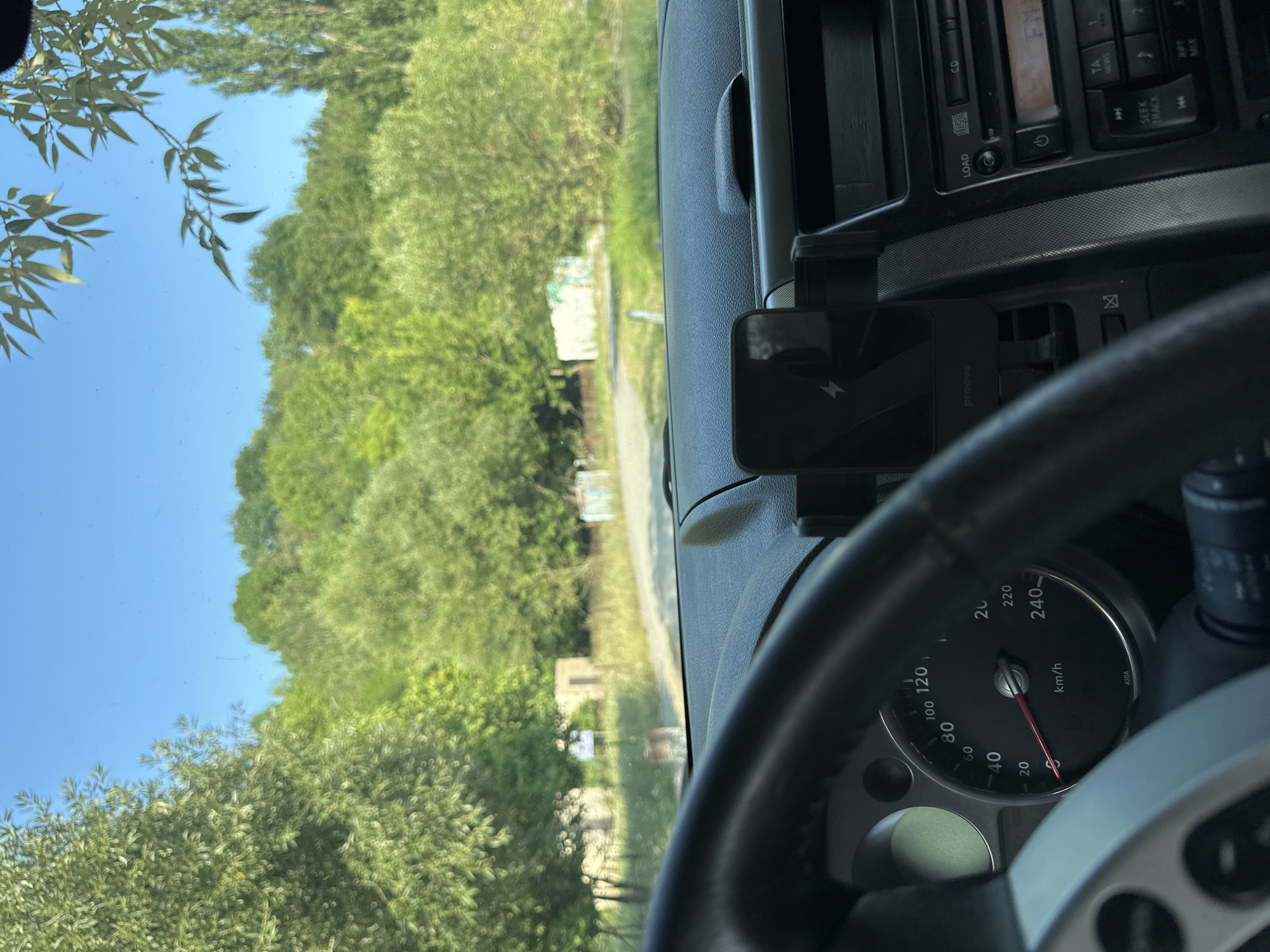 The image is taken from inside a car looking out through the windshield on a sunny day. The foreground shows the car's dashboard, including a steering wheel, speedometer displaying 40 km/h, and a phone mounted on the dashboard. The background depicts a rural landscape with lush green trees, a dirt road, and two small, weathered structures. The sky is clear blue. The car appears to be parked or driving slowly on a road in a rural area, possibly on the outskirts of Makiv, Ukraine, given the context. There are no people visible in the image.