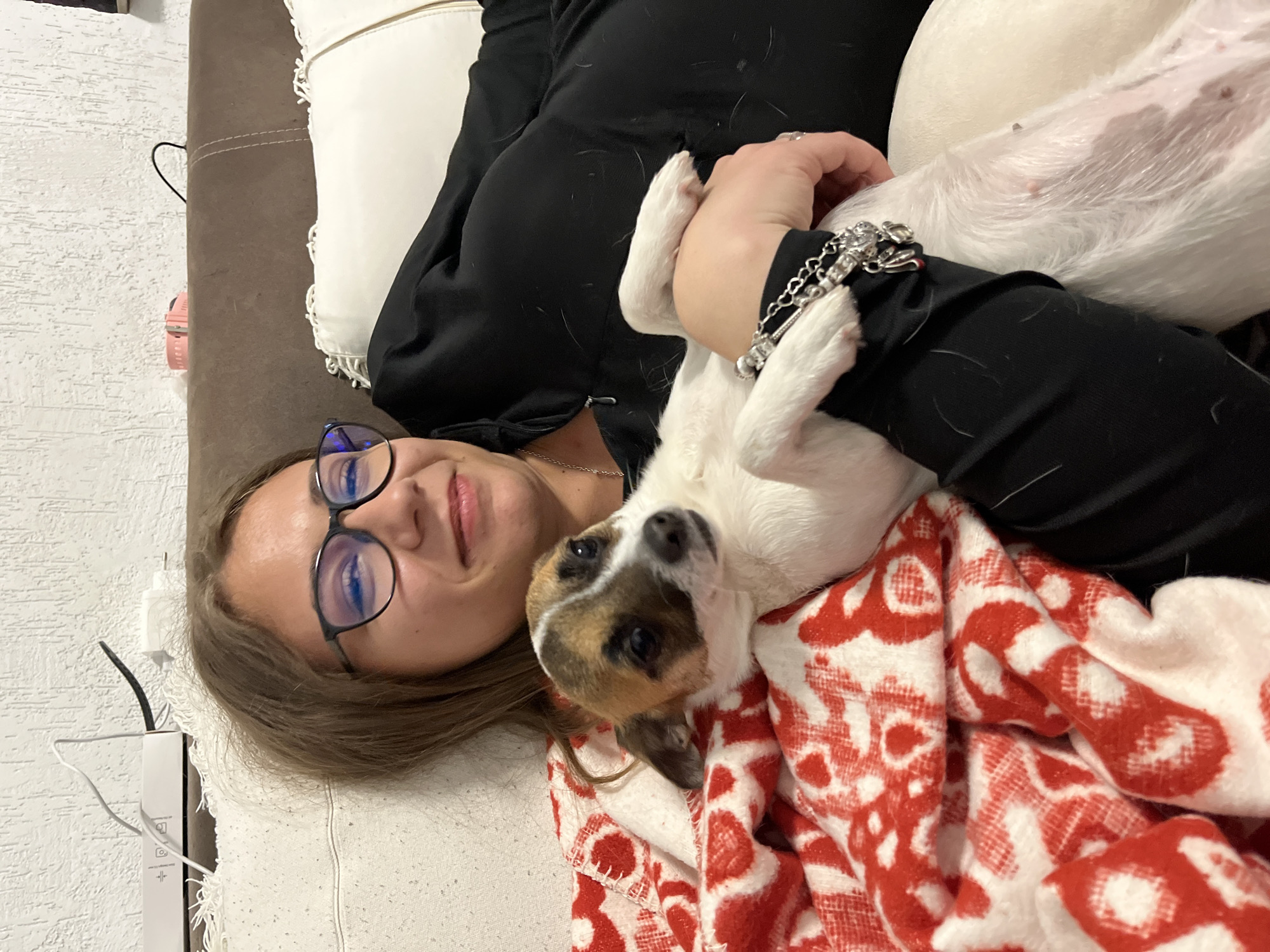 The image captures a woman lying on a light-colored surface, likely a sofa or bed, covered with a brown material and decorated with white pillows. She is wearing glasses and a black hooded top, and a silver bracelet is visible on her wrist. A small dog with white and brown fur is lying on top of her, partly covered by a red and white patterned blanket. The background features a white textured wall. There is no information in the image indicating that this is in the city of Karavelovo, Bulgaria.