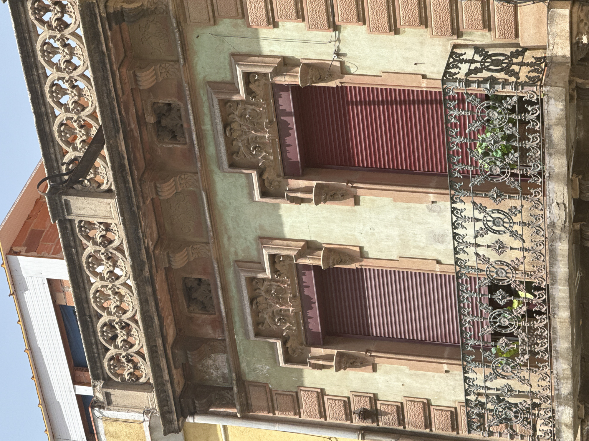 The image showcases the ornate architectural details of a building's facade in Barcelona, Spain. The focus is on decorative elements, including intricate wrought-iron railings and carved stonework adorning the windows and balconies. Two windows, each with reddish-brown shutters, are visible, framed by elaborate, carved stone surrounds featuring floral and scrolling motifs.
A decorative wrought-iron balcony, with circular patterns and swirling vine-like designs, extends across the right side of the frame. Through the balcony, glimpses of green plants can be seen, suggesting a residential or decorative use of the space. Above the windows, a row of rectangular, textured panels adds further architectural interest to the building's upper section.
The building's exterior is painted in muted tones, with a pale green plaster dominating the wall space around the windows. The stonework and decorative elements appear to be made of a reddish-brown material, possibly terracotta or sandstone, which contrasts with the plaster. The lighting suggests it is daytime, with sunlight casting shadows on the facade, highlighting the three-dimensional nature of the carvings and ironwork. The sky is visible on the left, appearing clear and blue. The overall impression is one of historical architecture with a high level of craftsmanship. No people or specific events are depicted, making the image a study of the building's aesthetic features.