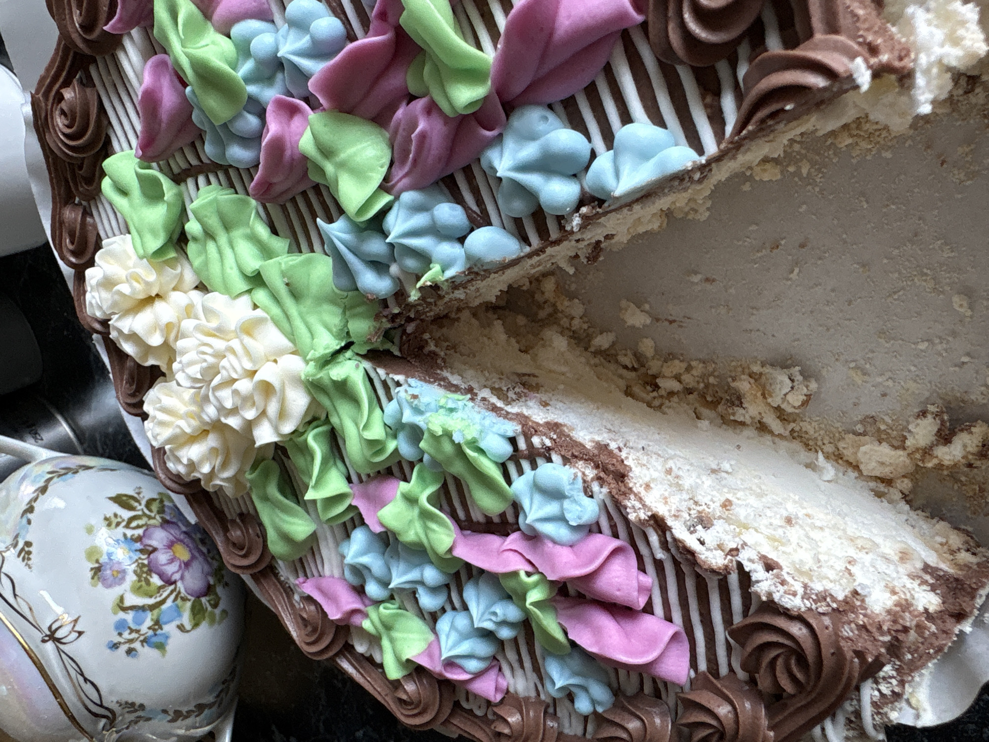 This close-up, high-angle image captures a beautifully decorated cake with a slice already removed, indicating it is in the process of being served or consumed. The scene is set indoors, in Hatne, Ukraine.
The cake is adorned with an array of pastel-colored frosting elements on its top surface. These include intricate light green leaves, delicate light blue rosettes, and ruffled pink embellishments, alongside clusters of cream-colored floral designs. The cake's outer edge features a rich chocolate frosting piped in decorative swirls, complemented by crisp white vertical stripes that extend down its side. The cut section of the cake reveals its internal structure, composed of multiple creamy layers and visible crumbs, suggesting a rich texture.
To the left of the cake, a portion of an ornate porcelain teapot or cup is visible. It is decorated with an elegant floral pattern featuring purple and blue flowers and green leaves, accented with subtle gold trim. The background is dark and out of focus, allowing the detailed cake and tableware to stand out. There are no people visible, nor any discernible text. The image does not provide cues regarding the time of day or weather. The overall impression is one of an inviting and artfully prepared dessert.