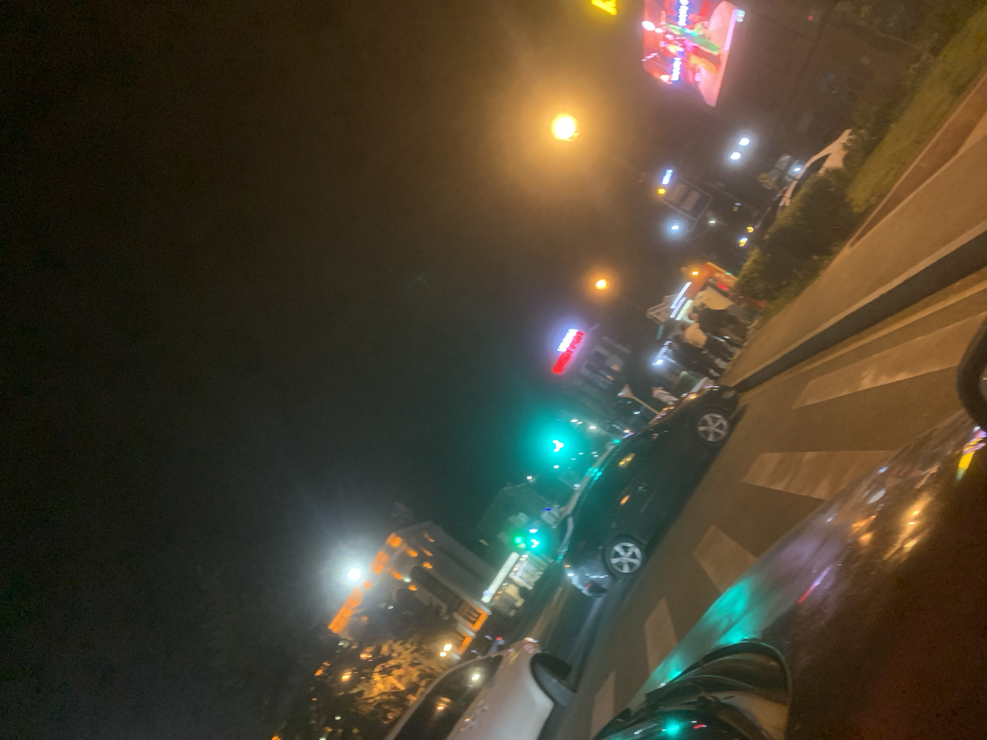 This image captures a night scene on a city street in Gyumri, Armenia, taken from within a vehicle. In the immediate foreground, a dark vehicle surface, likely a dashboard or hood, reflects a prominent green light. The street features a clearly marked white pedestrian crosswalk.
Midground elements include a dark-colored sedan with visible alloy wheels, stopped or moving slowly, and a partial view of a white car on the left, both oriented away from the camera. An overhead green traffic light signals active vehicular movement. To the right of the road, a sidewalk runs parallel to a narrow, grassy median strip, where a group of several pedestrians is visible walking.
The background features various urban elements under the dark night sky. On the left, multi-story buildings are illuminated with warm orange and yellow lights, showcasing architectural details that suggest a classical or historic style, possibly including arched windows or cornices. A bright, diffused light source, potentially the moon or a powerful streetlamp, is visible above these buildings. On the right, a large digital billboard emits vibrant, colorful light, displaying animated figures, likely an advertisement. Several streetlights cast orange and yellow glows throughout the scene. A red sign with white text is partially visible on a building on the left, and a smaller sign, possibly a bus stop, stands near the pedestrians on the right. The scene depicts a lively urban environment at night, bustling with both vehicle and pedestrian traffic.