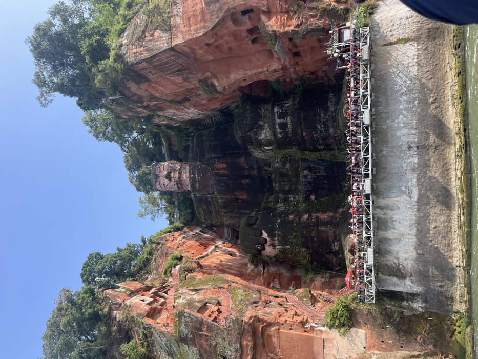 The image displays the monumental Leshan Giant Buddha, a colossal stone statue carved into a reddish-brown sandstone cliff face, located in Shizhong, Leshan, Sichuan Province, China. The seated Buddha, facing forward, dominates the central and upper portions of the scene, with its head and upper torso exhibiting weathered stone tones, while the lower body and legs are extensively covered in dark green moss and mineral staining. Verdant trees and foliage crown the cliff edges surrounding the statue. To the left, a series of red-railed stairs and carved sections are visible on the cliff face. At the base of the Buddha, a long, elevated metal viewing platform, supported by a vertical grey rock wall, extends horizontally above a body of greenish-brown water. Approximately 100-150 individuals, appearing as tourists, are densely packed along the platform, observing the statue. Small structures, including red umbrellas, are positioned on the platform. The upper sky is clear and bright blue, indicating daylight conditions.