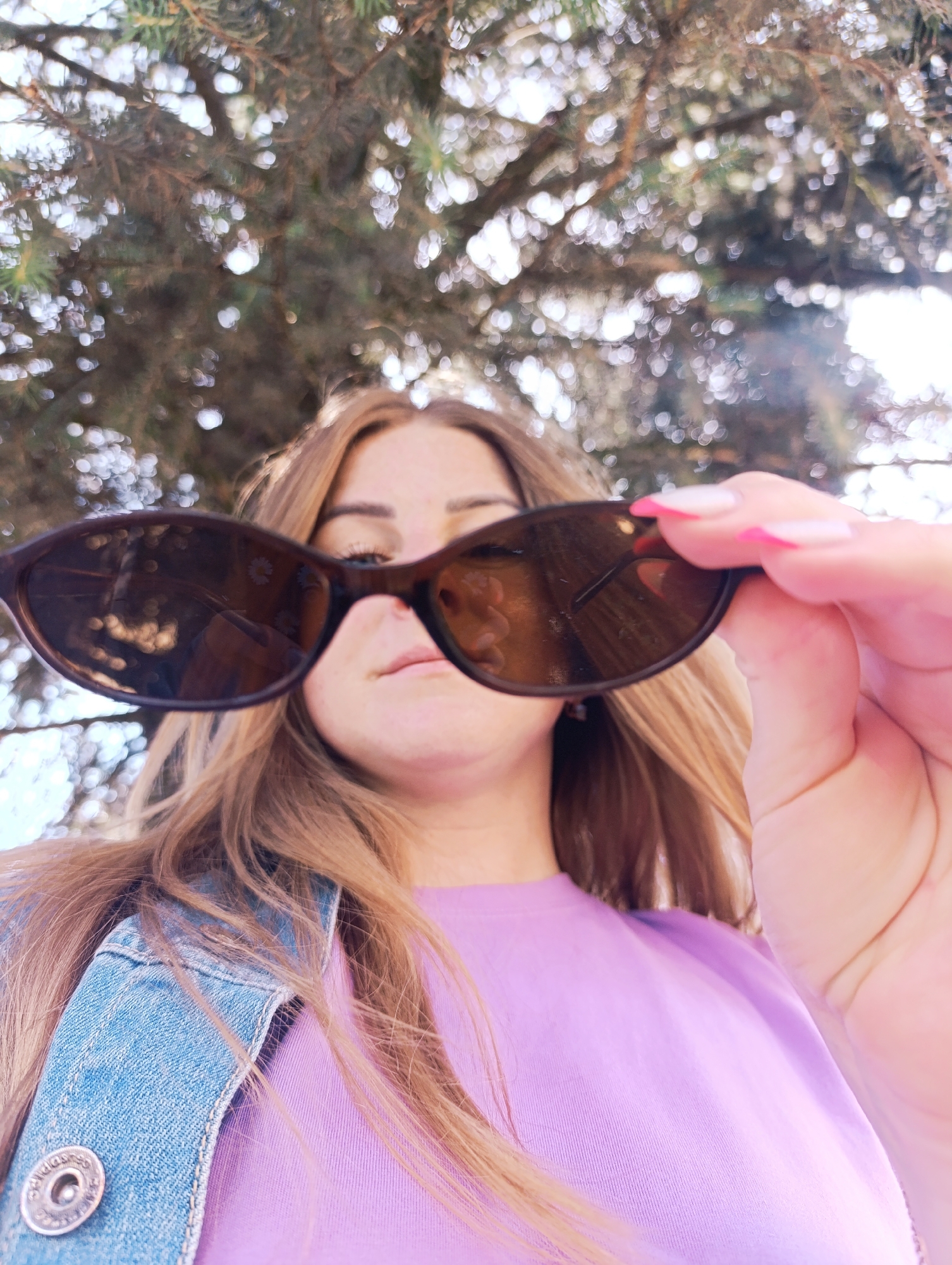A low-angle, medium shot shows a woman holding a pair of dark-lensed sunglasses in front of her face, obscuring her eyes. She has long, light brown hair that is slightly blurred and appears to be outdoors, with out-of-focus branches of a coniferous tree forming a bokeh effect in the background. The woman is wearing a light purple t-shirt and a blue denim jacket with a silver button visible on the lapel. Her right hand, with bright pink-tipped French manicured nails, is holding the sunglasses up. The lighting suggests it is daytime, possibly sunny.