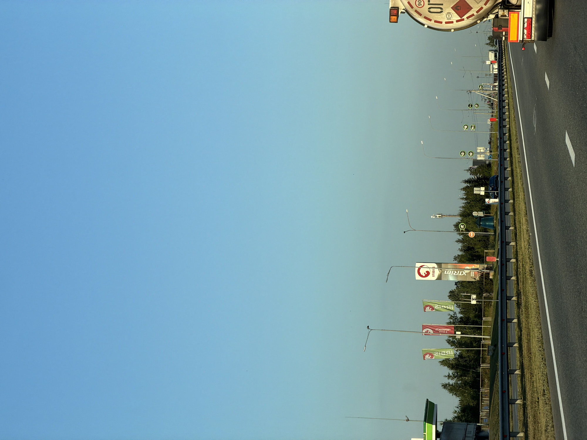 The image shows a highway scene in Kulaevo, Russia, under a clear blue sky. The foreground features a paved road with white lane markings, bordered by a metal guardrail and a grassy shoulder. To the left, a gas station canopy with green trim is visible, along with several tall flagpoles displaying flags with red and white logos. Further down the road, a tanker truck, labeled with hazard symbols and the number 90, is in the right lane. Other vehicles and streetlights are sparsely visible in the distance. The background consists of a line of trees.